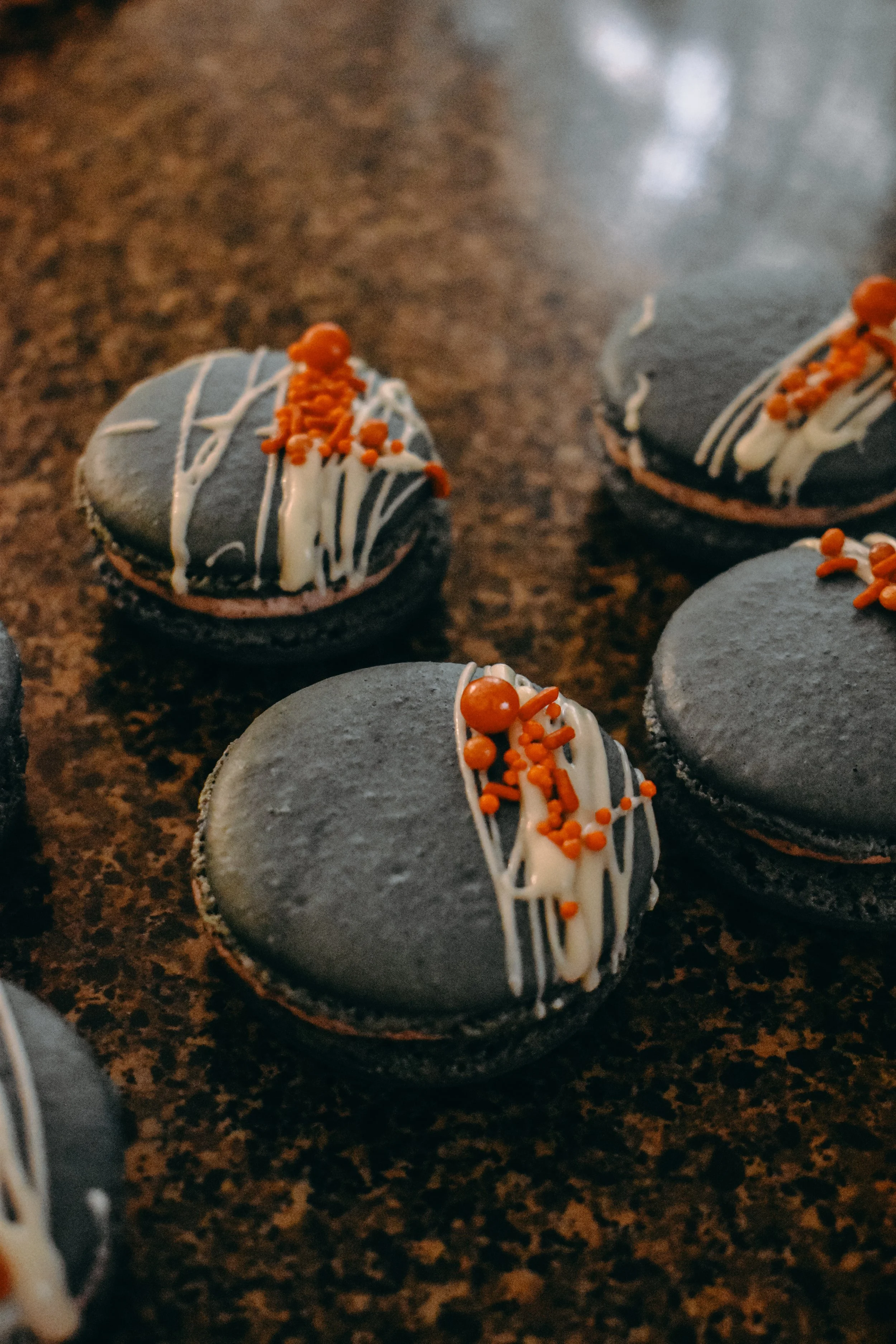 Close-up of black cookies decorated with white chocolate drizzle and orange sprinkles on a dark brown marble surface.