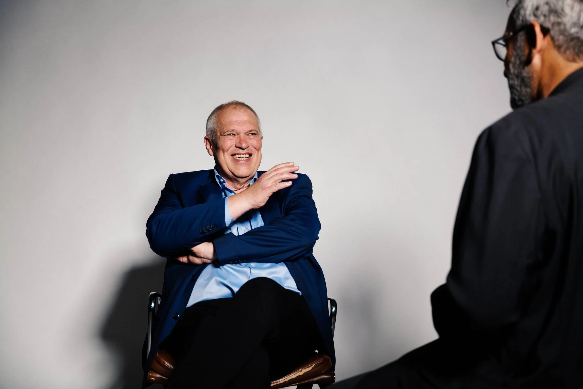 Martin Wegner in einem Gespräch. Der Mann auf der linken Seite lächelt und ist in einem blauen Anzug. Der andere Mann auf der rechten Seite trägt schwarze Kleidung und Brille. Sie sitzen vor einer weißen Wand.