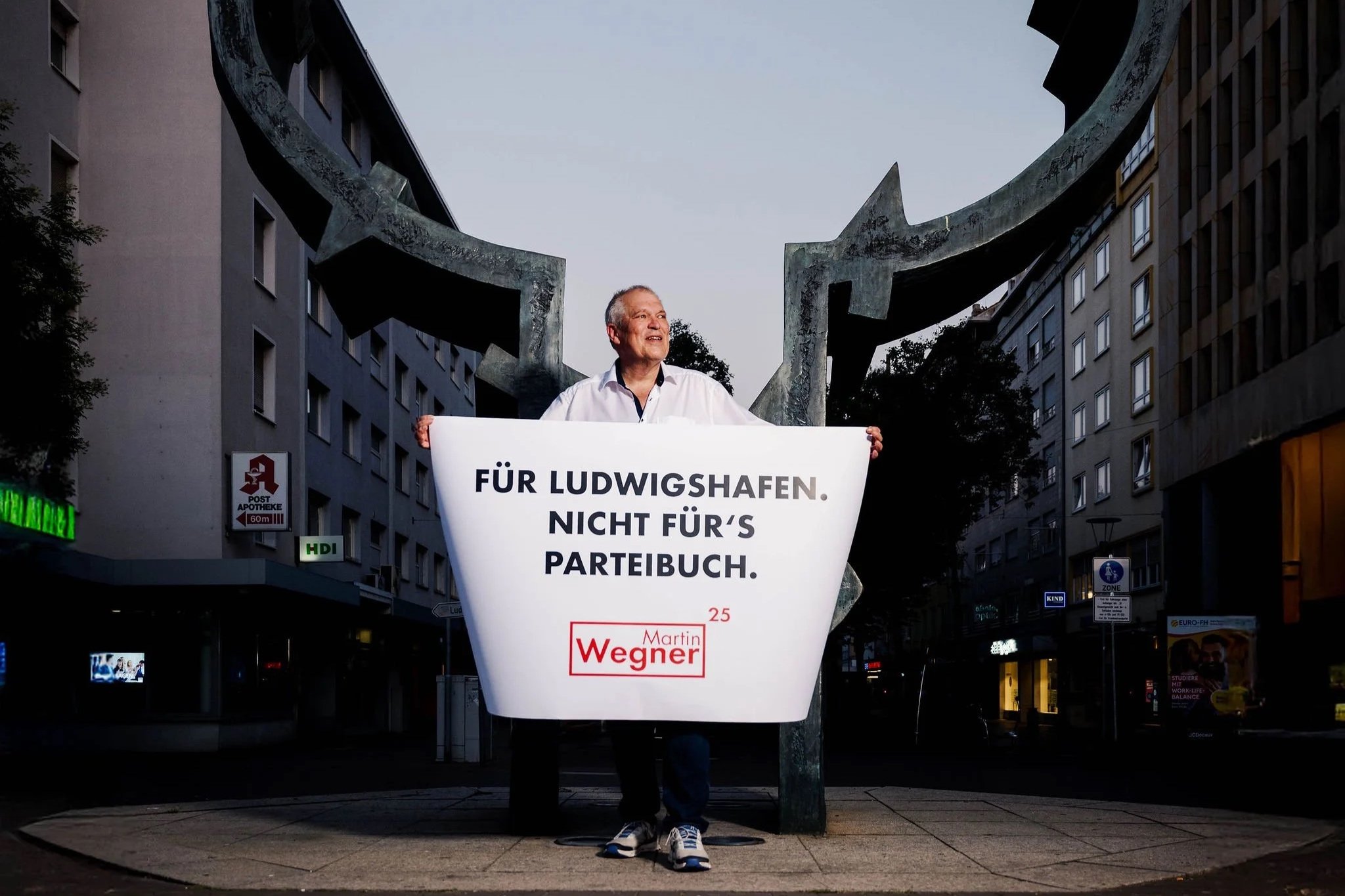 Martin Wegner Oberbürgermeisterkandidat für  Ludwigshafen  steht in der  Stadt bei Dämmerung und hält ein Plakat, das für Ludwigshafen ist, aber gegen ein Parteibuch protestiert. Im Hintergrund sind moderne Gebäude und eine Skulptur sichtbar.