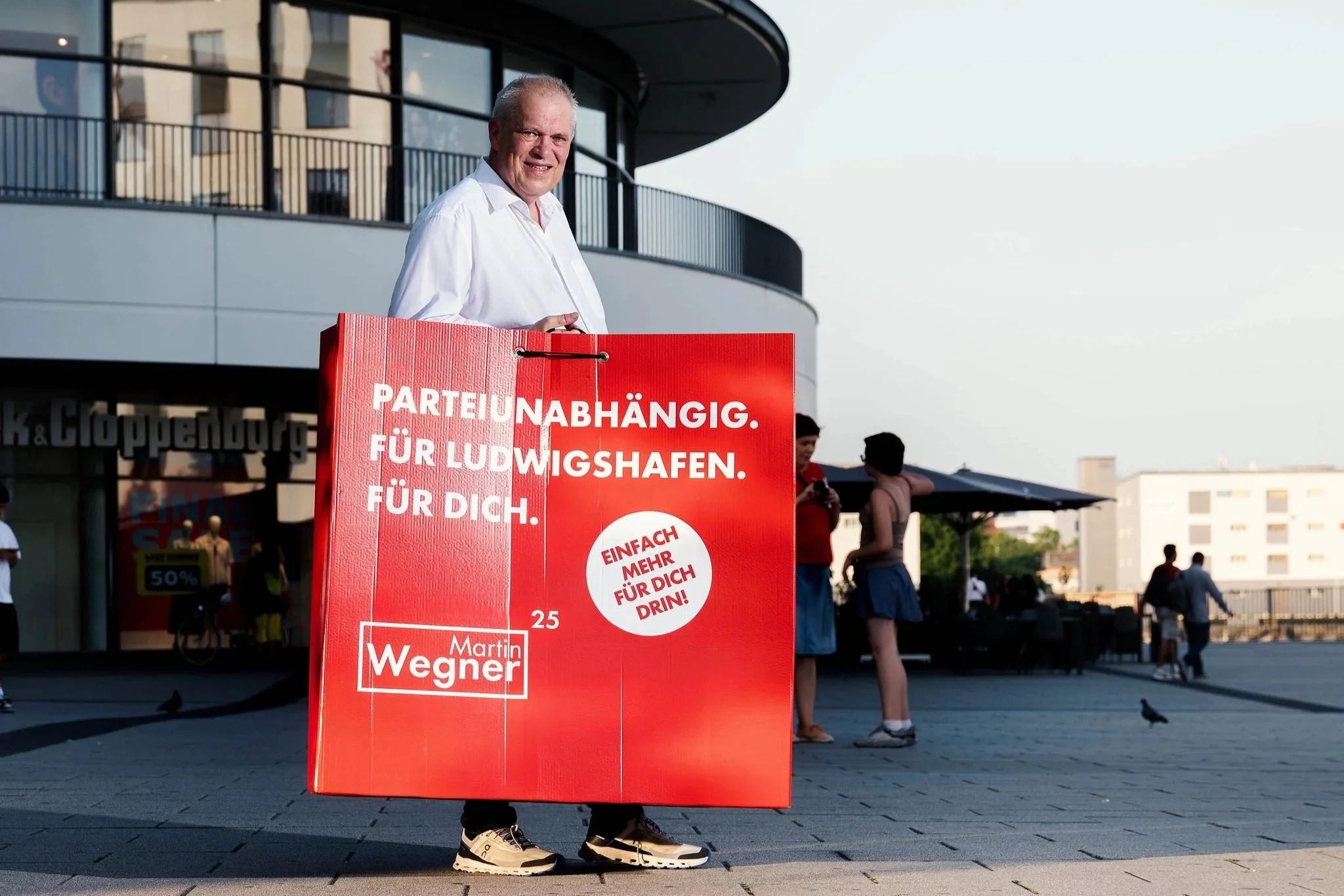 Martin Wegner Oberbürgermeisterkandidat für  Ludwigshafen trägt eine große Einkaufstasche  mit Text auf Deutsch. Hintergrund zeigt junge Menschen, die sich unterhalten, vor einem modernen Gebäude an einem sonnigen Tag.