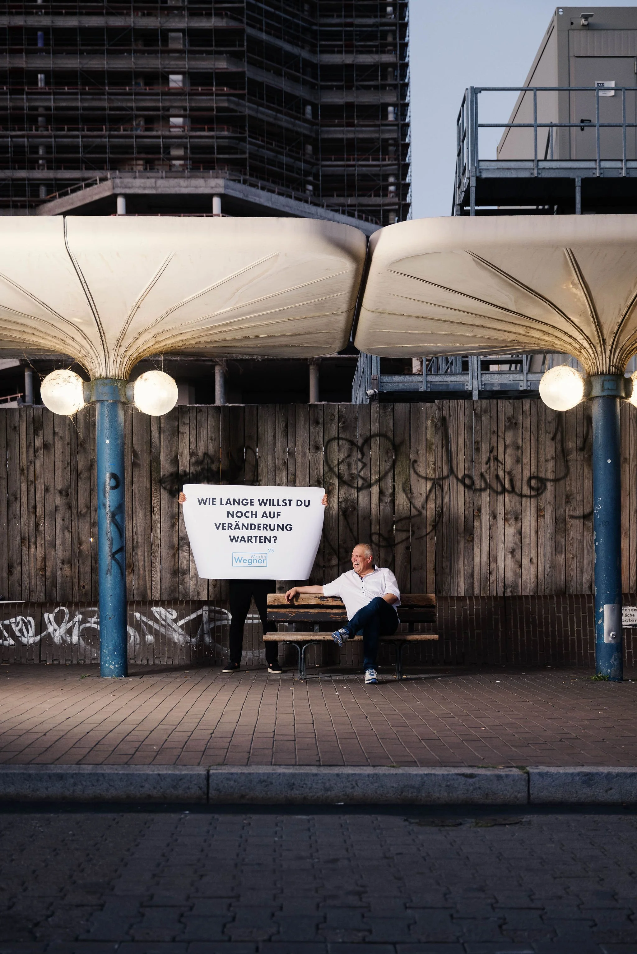 Martin Wegner Oberbürgermeisterkandidat für  Ludwigshafen  sitzt lachend auf einer Bank unter einer Bushaltestellen-Struktur mit zwei Lampen. Hinter ihm hängt ein Schild mit der Aufschrift "Wie lange willst du noch auf Veränderung warten?".