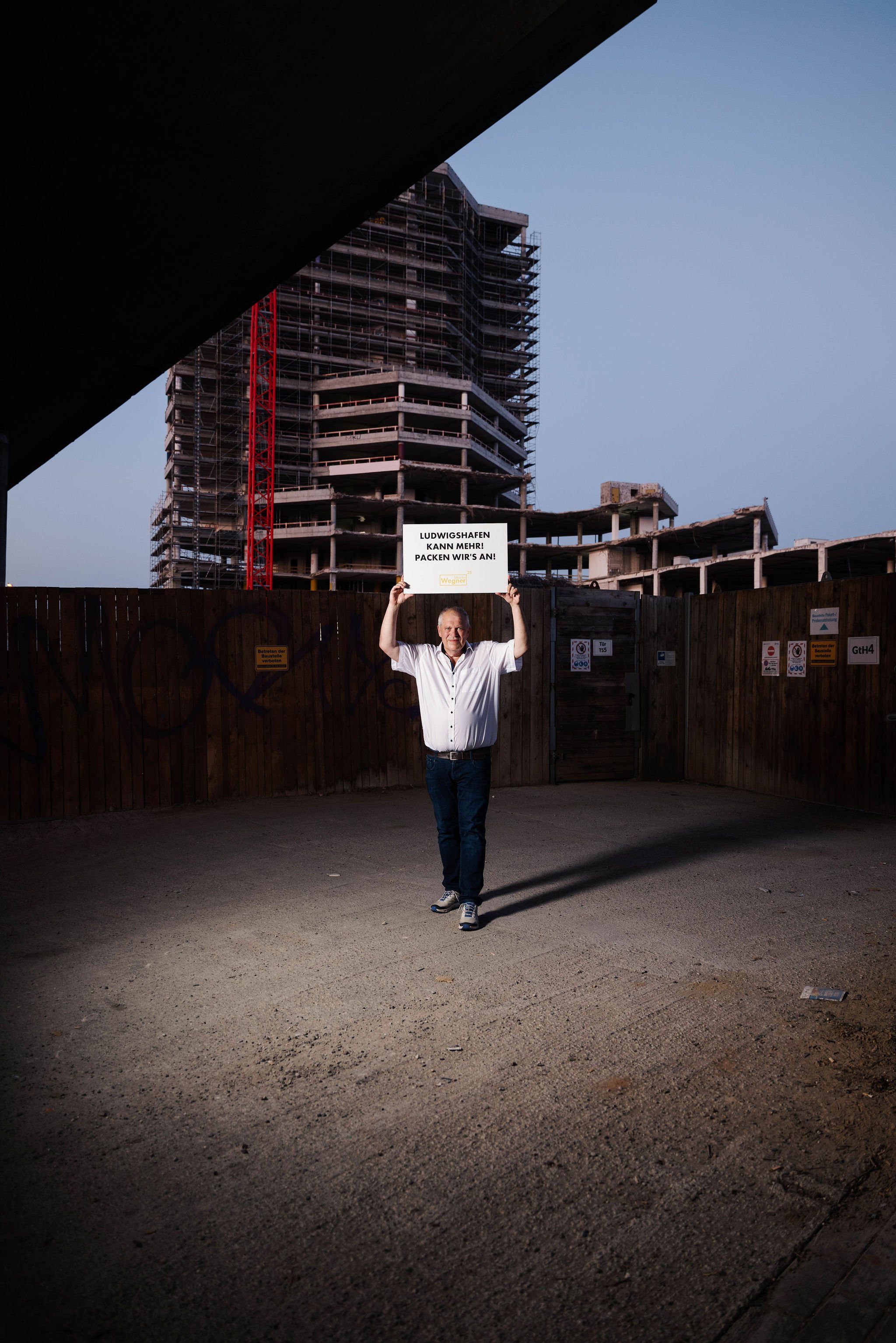 Martin Wegner Oberbürgermeisterkandidat für  Ludwigshafen  steht vor einer Bauzone, hält ein Schild hoch, auf dem steht 'Ludwigshafen kann mehr! Packen wir's an!'