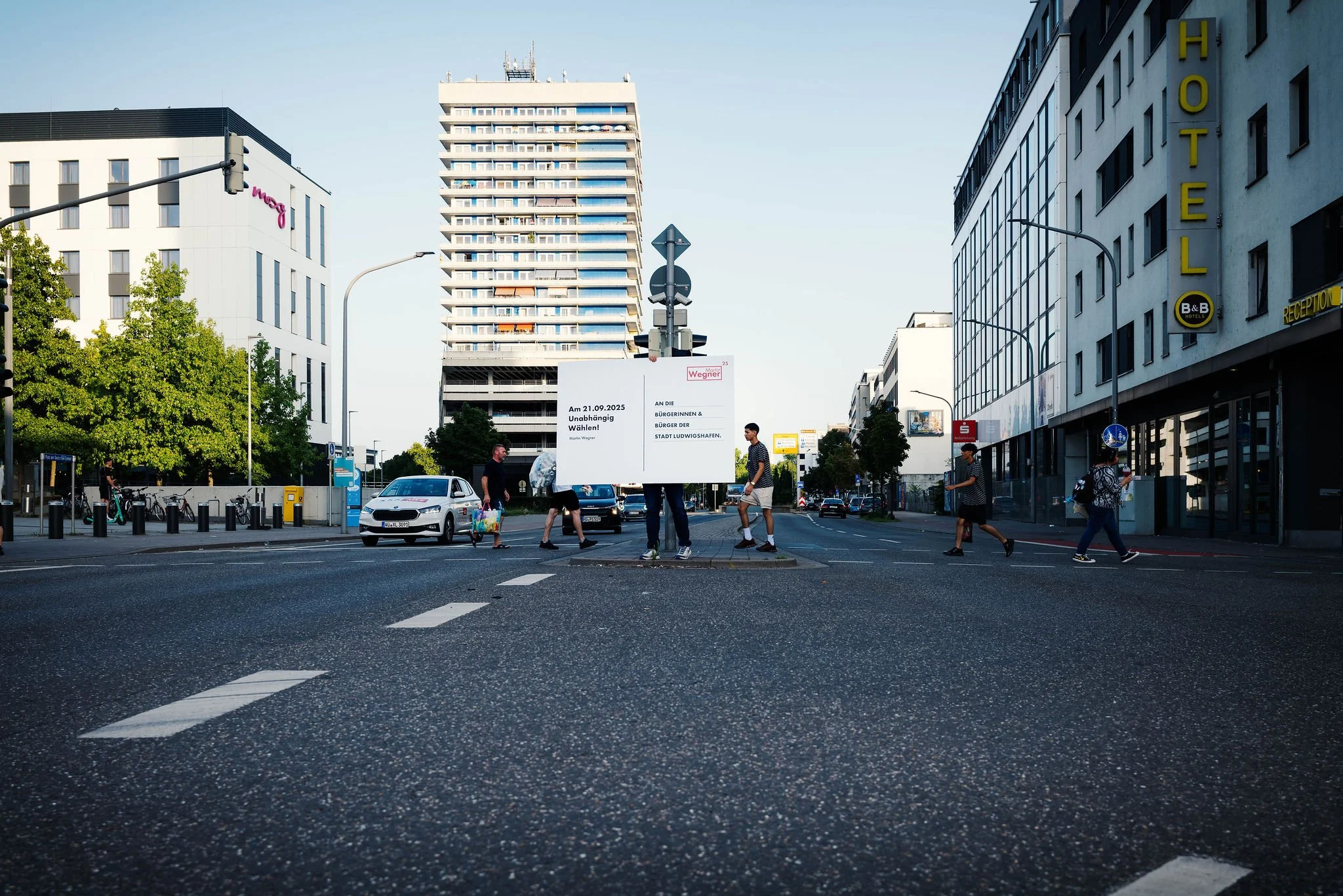 Straßenkreuzung in einer Stadt mit mehreren Fußgängern, die die Straße überqueren, einem großen Wahlplakat von Martin Wegner Oberbürgermeisterkandidat für  Ludwigshafen mit  modernen Gebäuden und Autos.