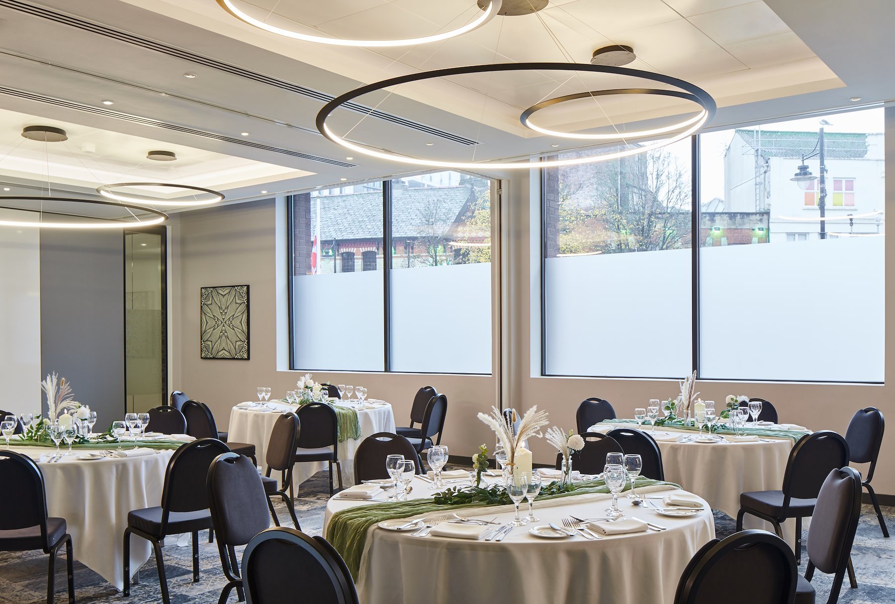 Elegant banquet room with round tables covered in white tablecloths, decorated with green runners and floral centerpieces, set for an event with glassware, plates, silverware, and chairs around each table. The room has large windows with frosted lower panels and modern circular ceiling lights.