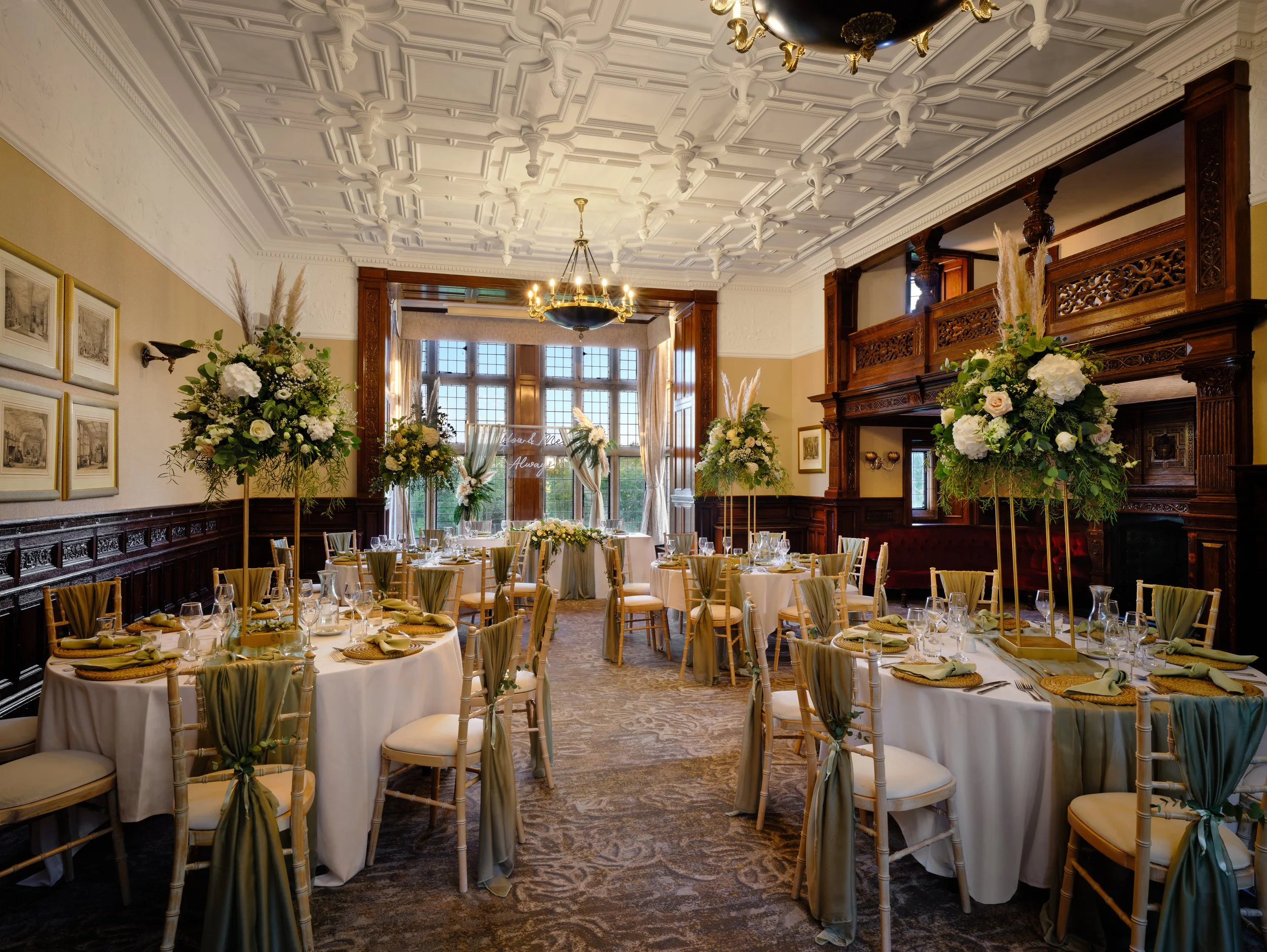 Elegant banquet hall decorated with floral centerpieces, round and rectangular tables set for a formal event, ornate woodwork, large windows, and detailed ceiling moldings.