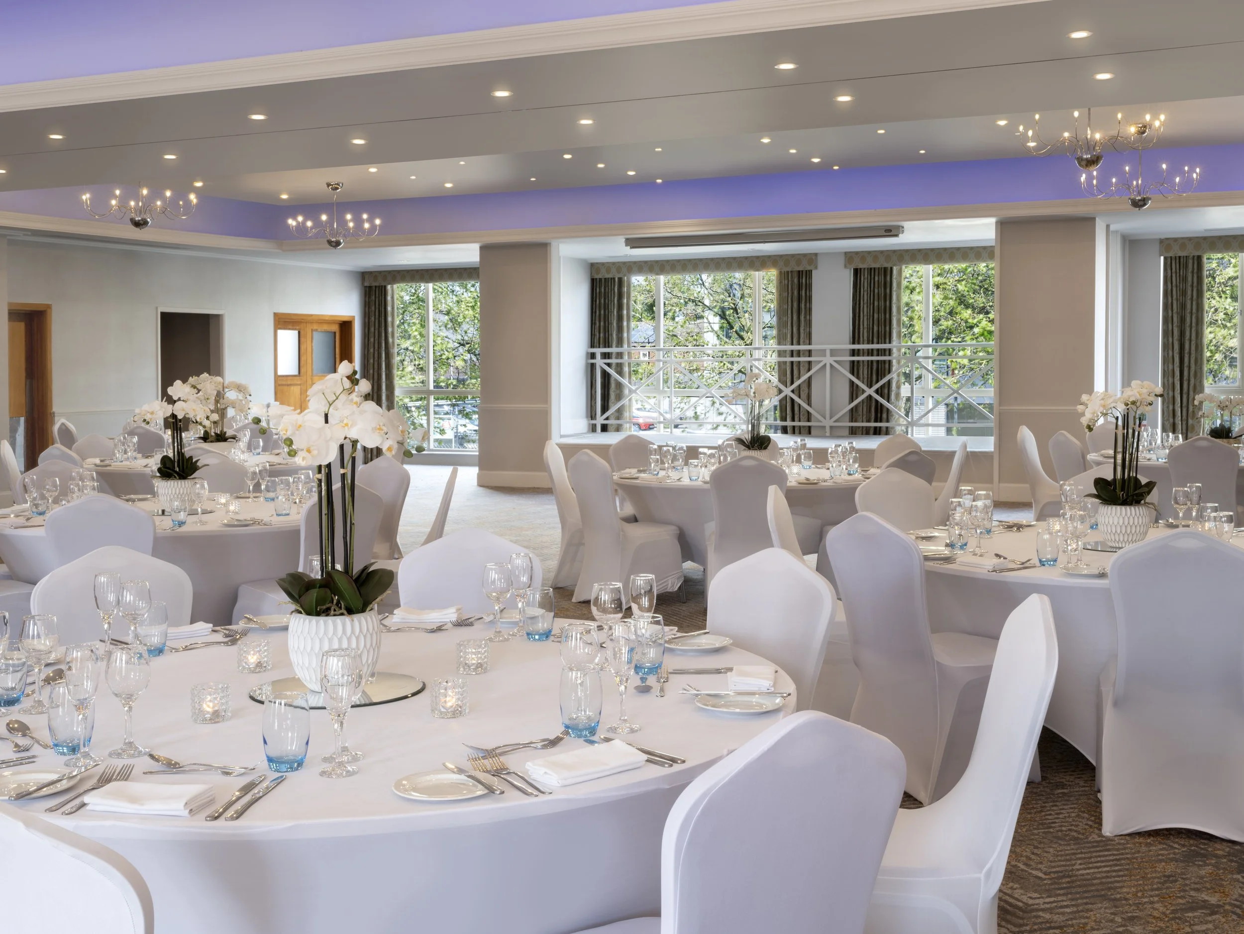 Reception or banquet hall with round tables covered with white tablecloths, set with glassware, silverware, and napkins, decorated with white orchids in vases, in front of large windows with patterned curtains and a view of trees outside.