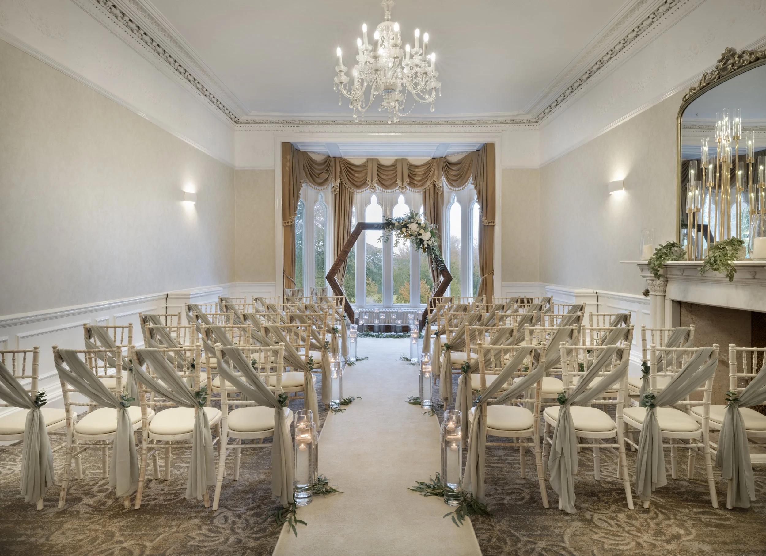 An elegant indoor wedding ceremony setup with rows of white chairs decorated with white fabric and green foliage, a white aisle, tall glass candles along the aisle, and a floral arch at the front near large windows adorned with gold drapes. A crystal chandelier hangs from the ceiling in a sophisticated room with cream-colored walls and detailed ceiling moldings.