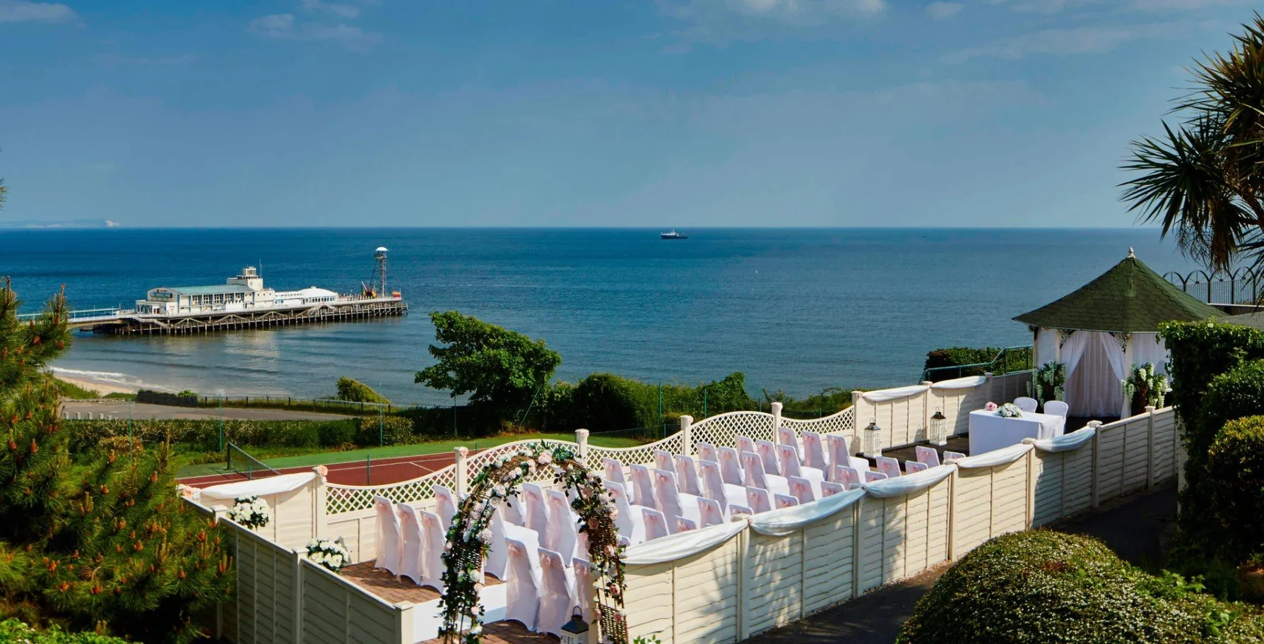 A wedding setup on a patio overlooking the ocean, with white chairs arranged in rows, a decorated arch, a gazebo, and a pier with a building extending over the water, under a clear blue sky.