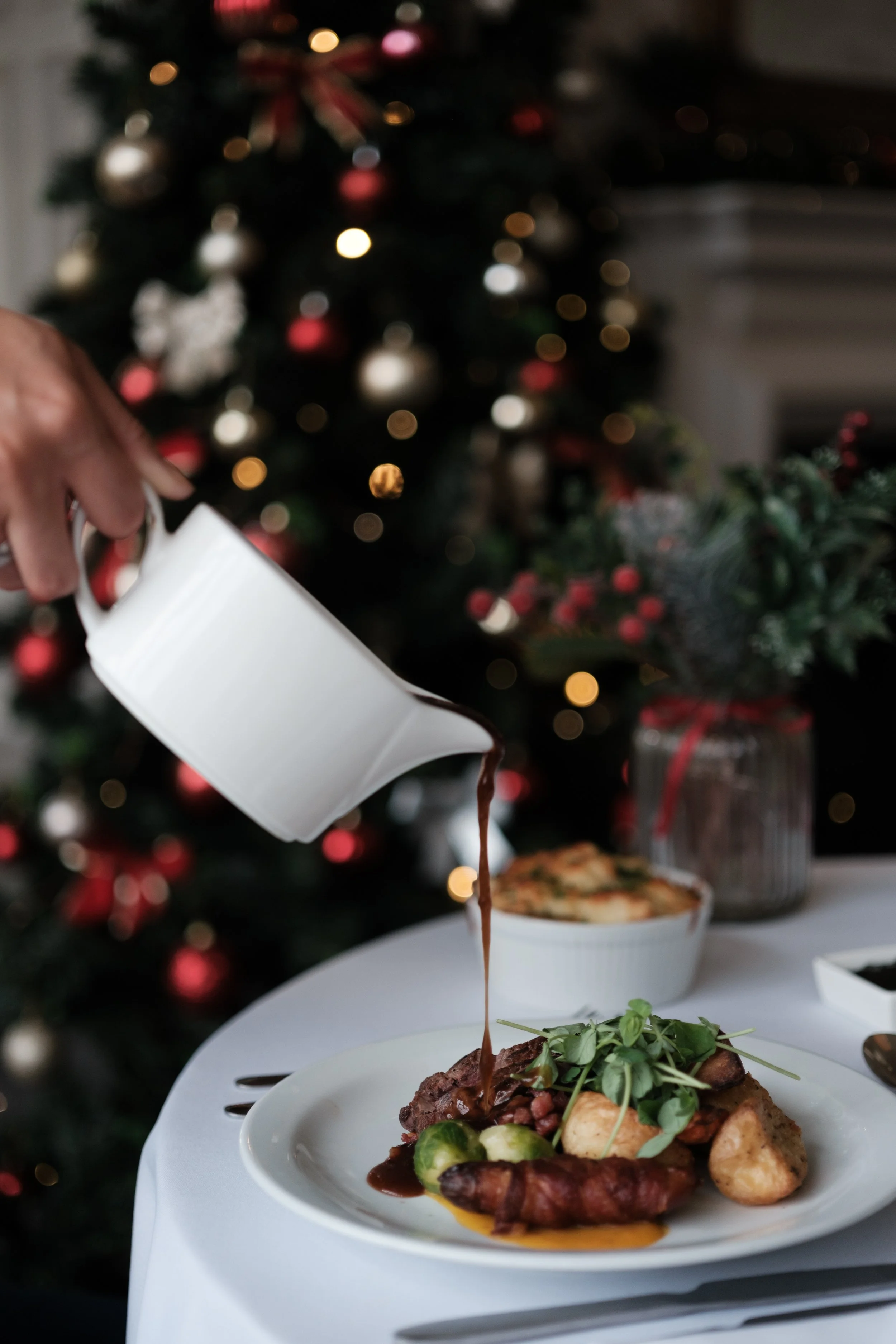 Pouring gravy over a plate of roast beef, potatoes, vegetables, and greens at a Christmas dinner table, with a decorated Christmas tree blurred in the background.