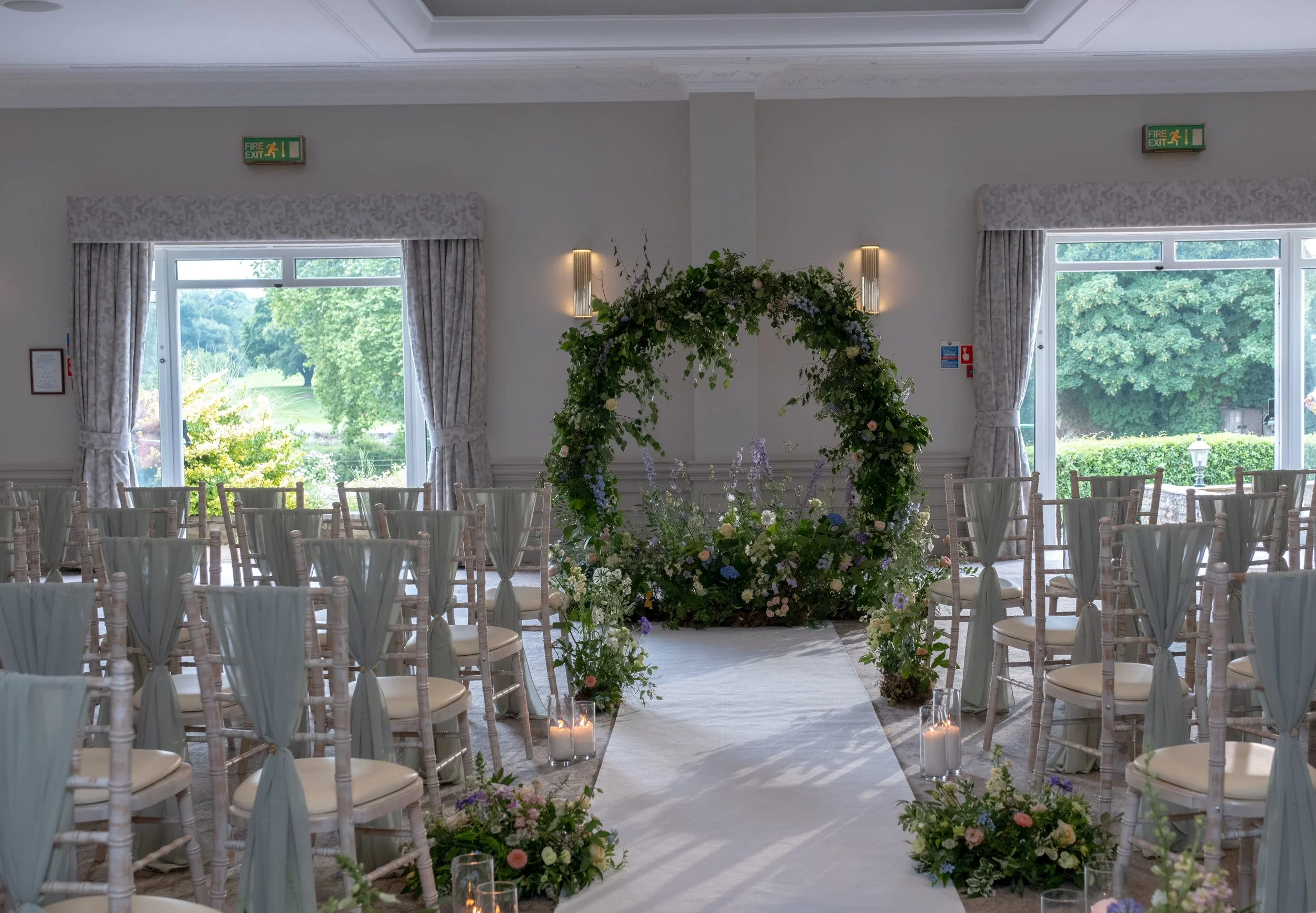 Wedding ceremony setup with arranged chairs, floral decor, and candles, near large windows showing greenery outside.