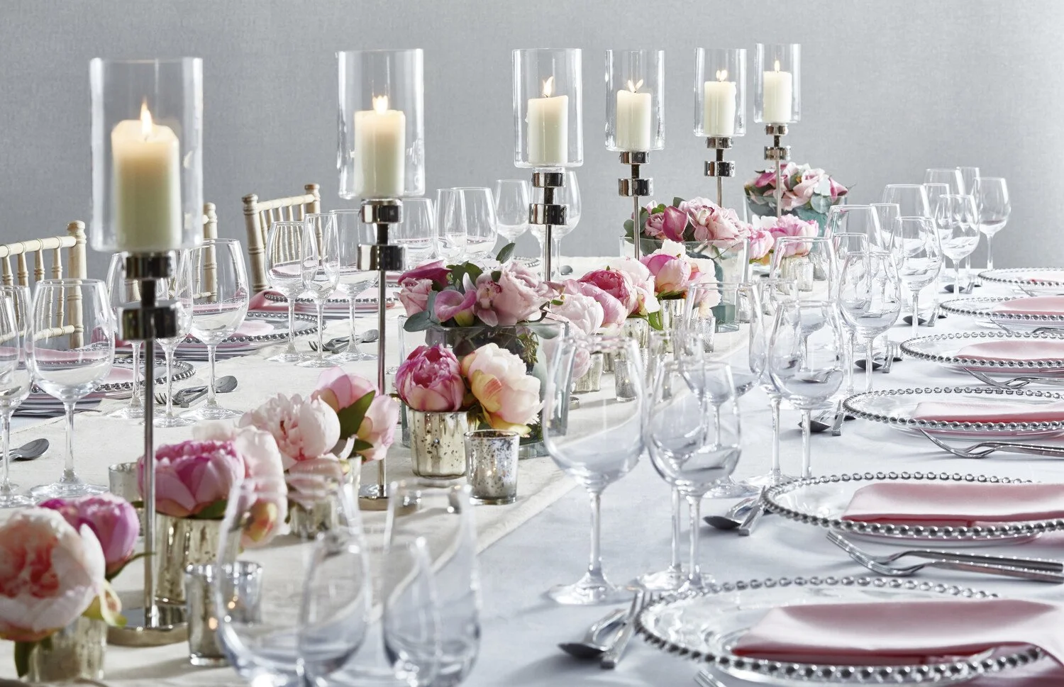 Elegant dining table set with pink peony and rose floral centerpieces, tall candles in glass holders, and clear glassware on a white tablecloth.