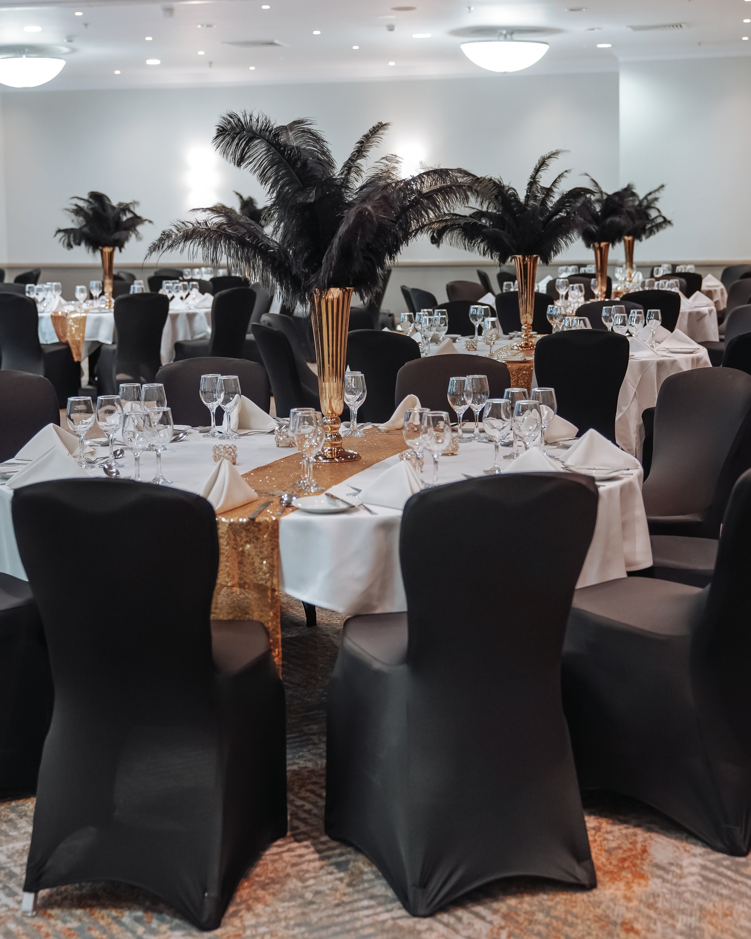 Elegant event setup with round tables draped in white tablecloths, each featuring glassware, napkins, and gold-colored table runners. Centerpieces consist of tall, gold vases filled with large black feathers. The room has soft ceiling lighting and a neutral-colored carpet.