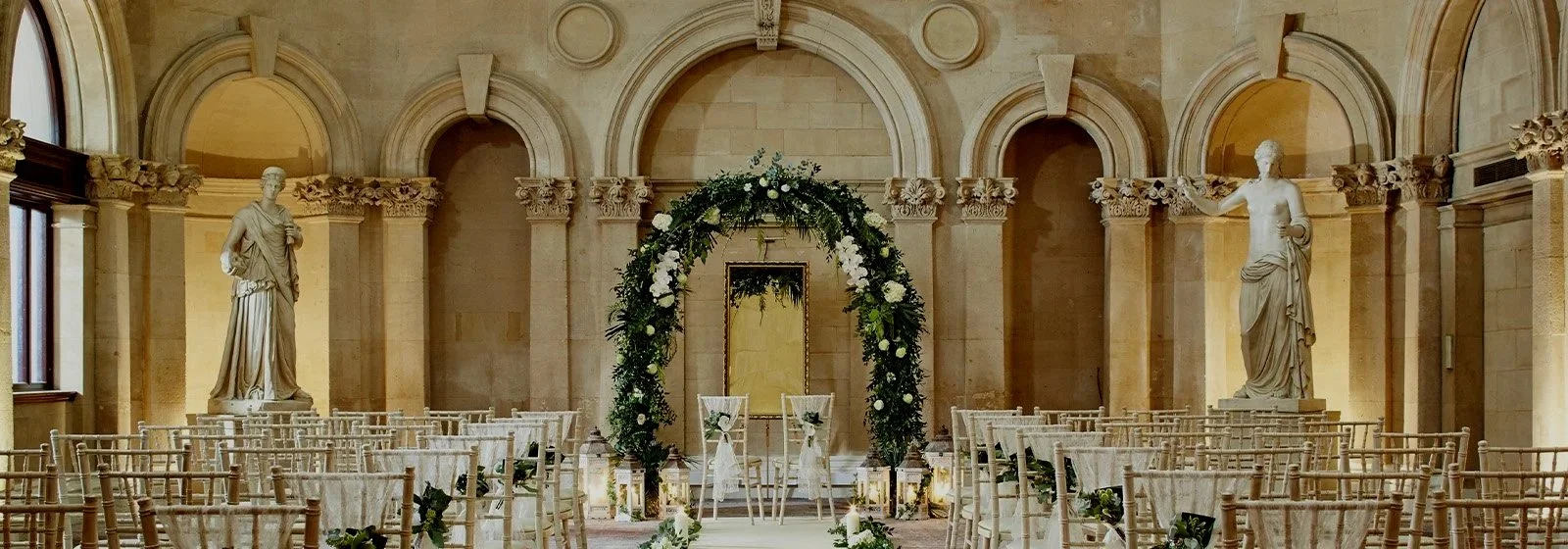 Indoor wedding ceremony setup with floral arch, rows of chairs, statues, and classical architecture.