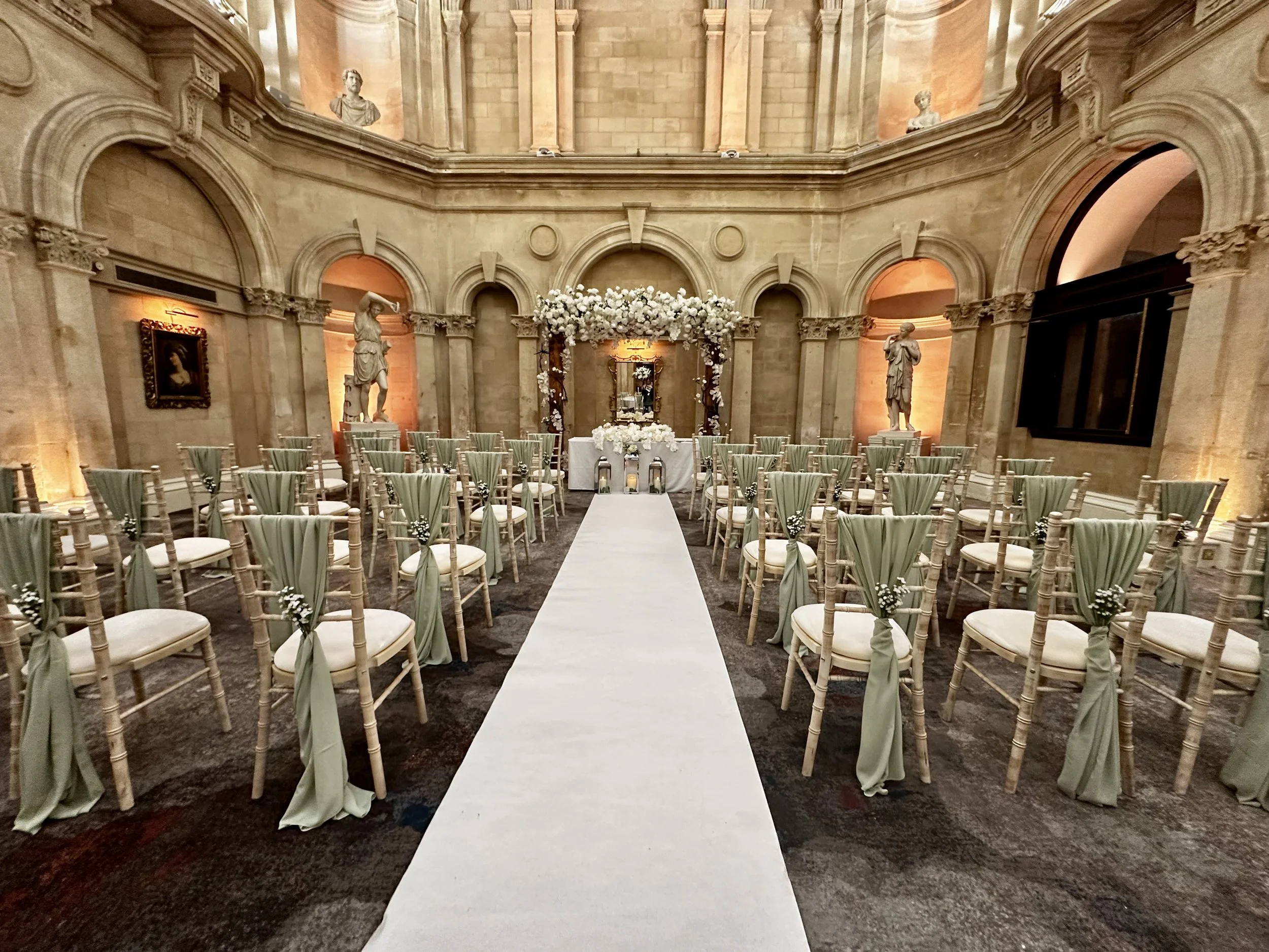 Elegant indoor wedding ceremony setup with rows of chairs decorated with green fabric and small white flowers, a white aisle runner, a floral arch at the altar, marble busts and framed artwork adorning the high stone walls.