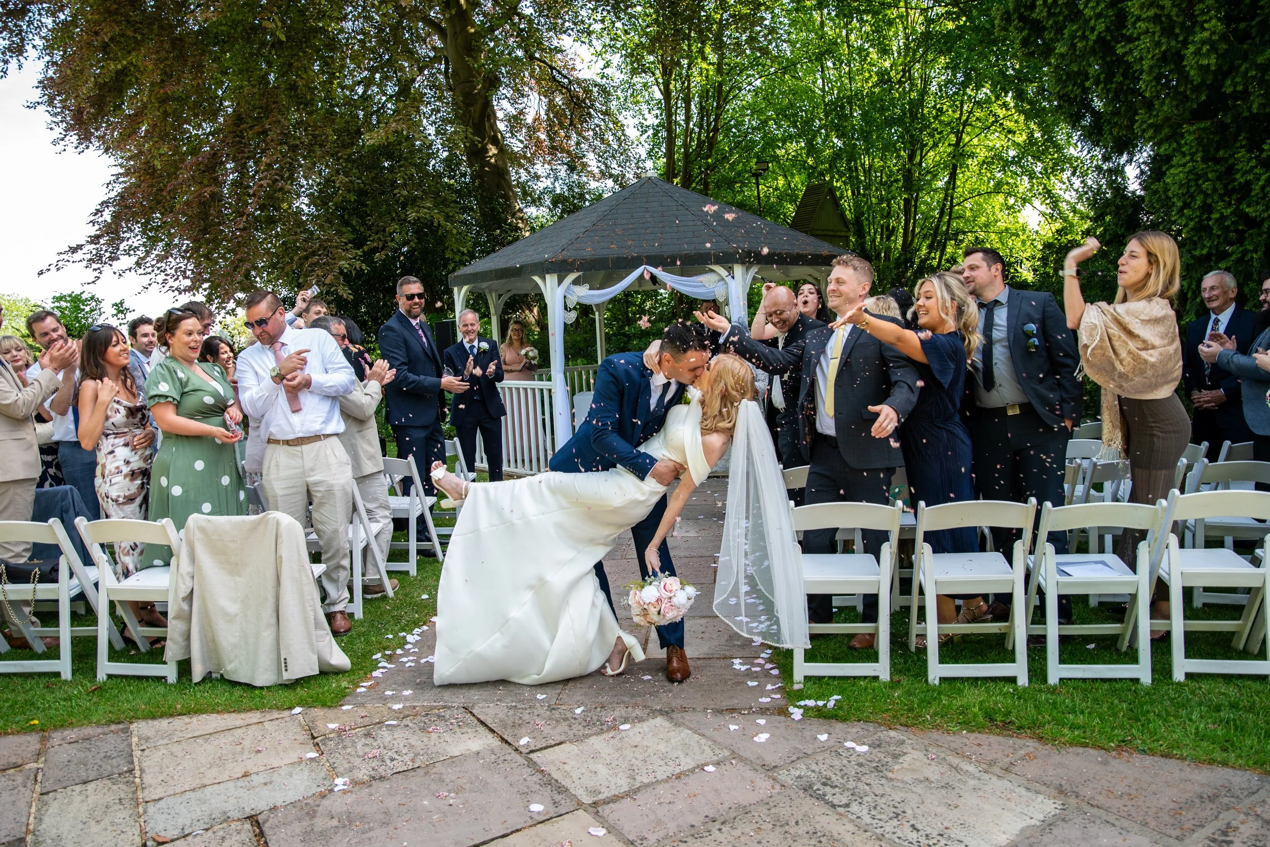 Wedding ceremony outdoors with the bride and groom, surrounded by guests, celebrating with confetti and smiling, under a gazebo and trees.