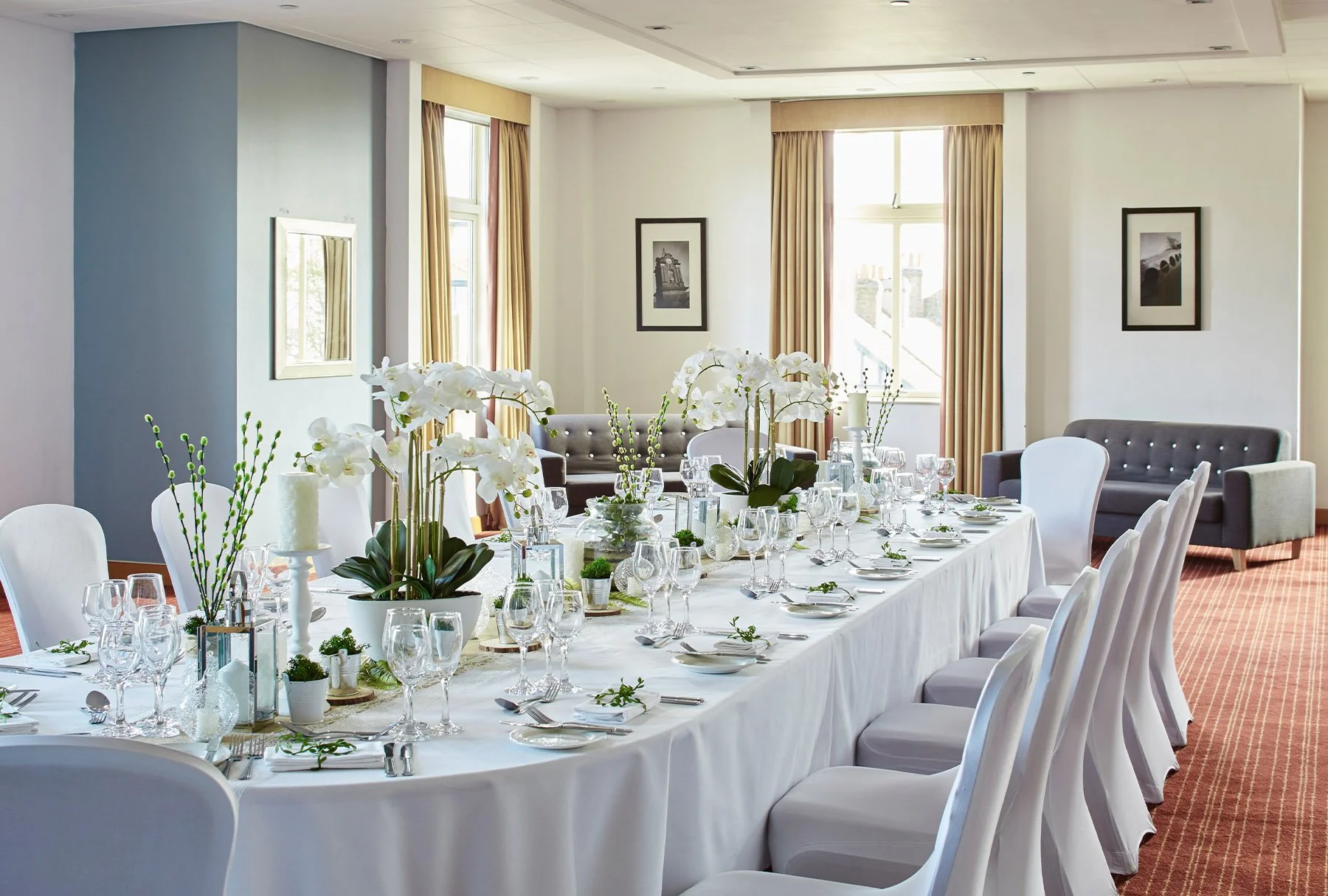 A well-decorated banquet table set for a formal event in a bright room. The table has white tablecloths, white chairs, and is decorated with white orchids, candles, and small potted plants. The room has large windows with beige curtains, framed black-and-white photographs on the walls, and a few sofas along the back wall.