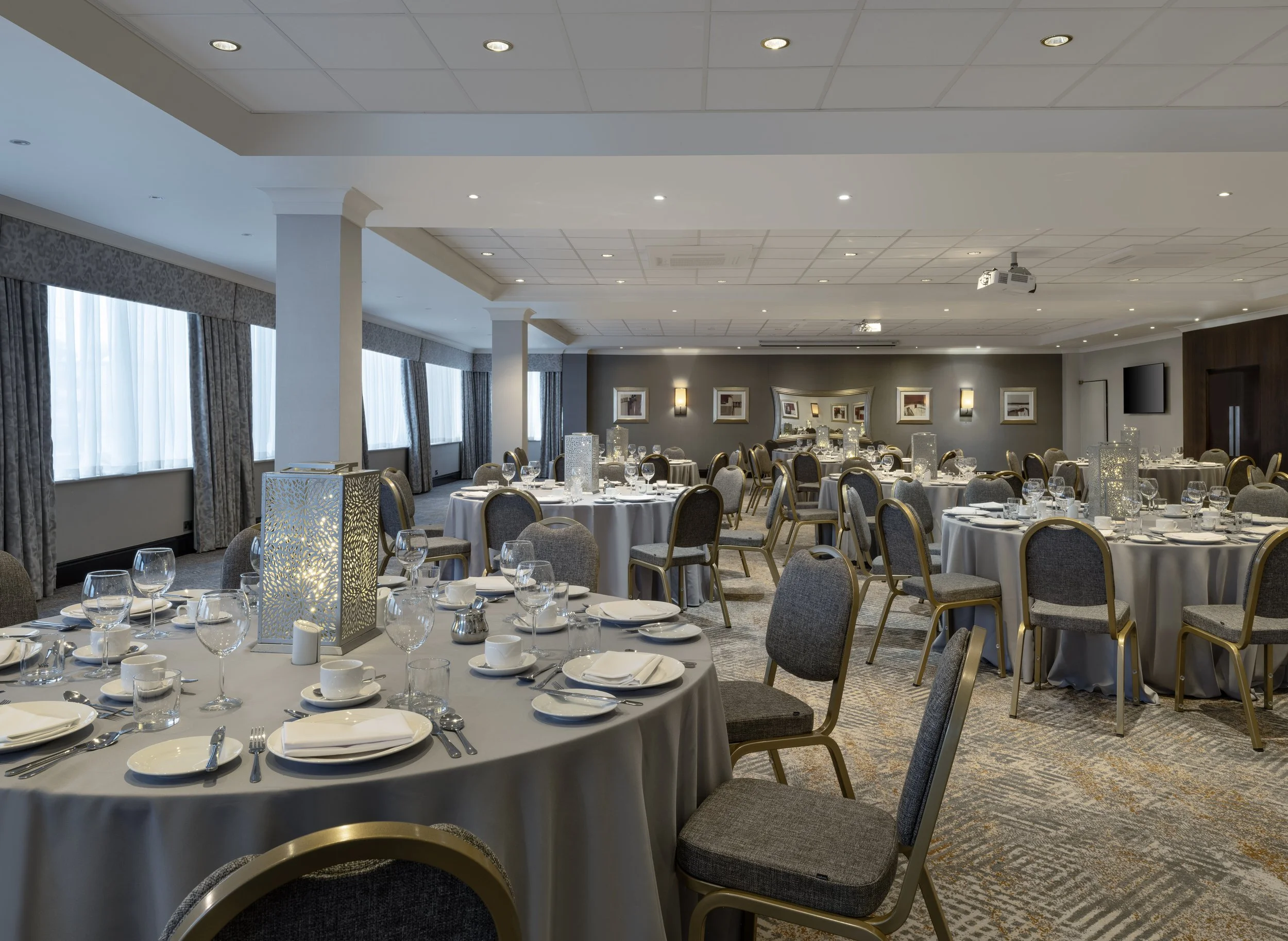 A banquet hall set up for a formal event with round tables covered in white tablecloths, neatly arranged silverware, glassware, and white napkins. The tables feature decorative candle holders and cylindrical lanterns. The room has large windows with patterned curtains, gray walls adorned with framed artwork, wall sconces, and a ceiling with recessed lighting and mounted projectors.
