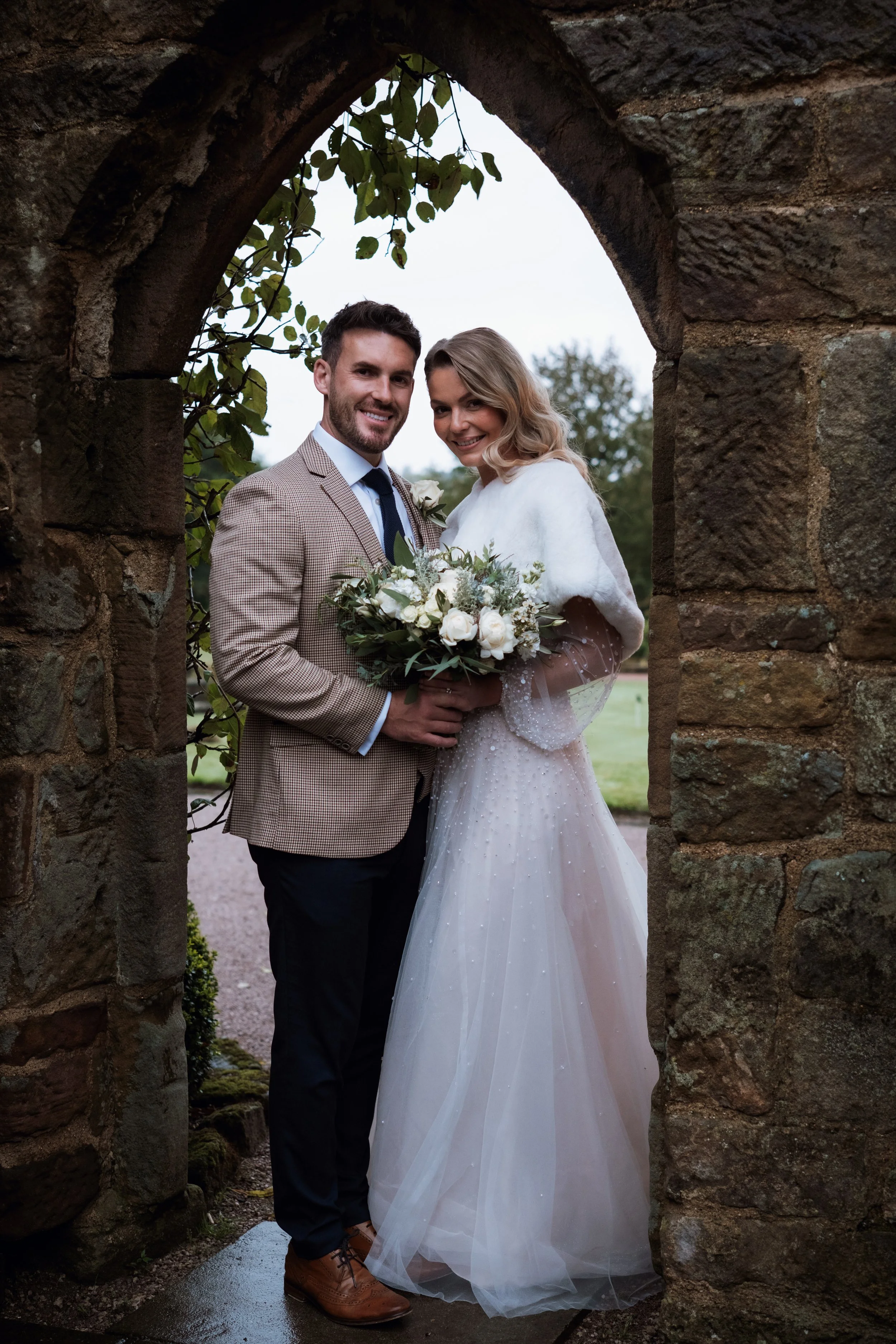 A bride and groom standing close together in a stone archway, smiling at the camera. The groom is wearing a checkered blazer, light blue shirt, and dark trousers, while the bride is wearing a white wedding gown and holding a bouquet of white roses and greenery.