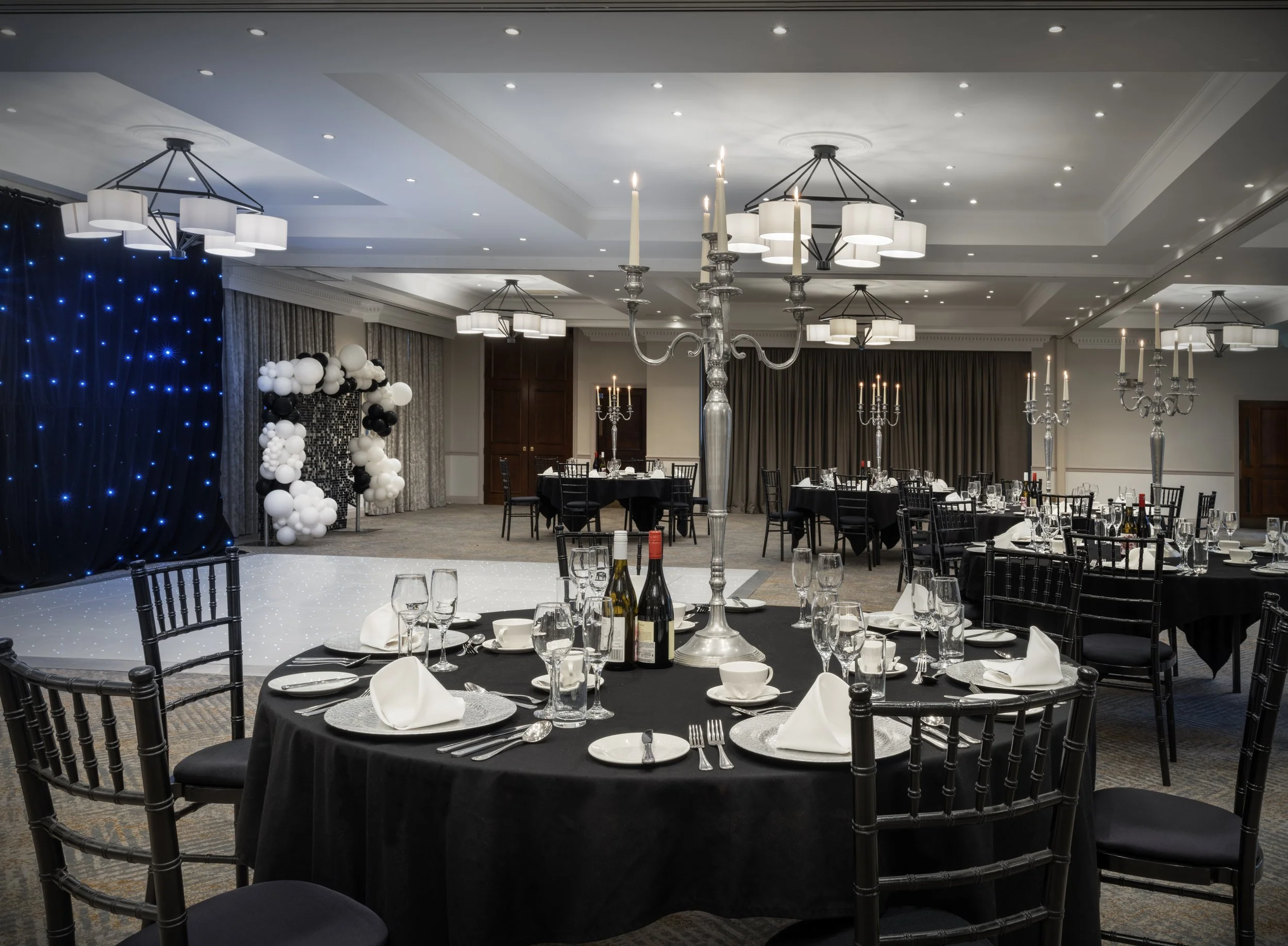 A banquet hall decorated for an event with round tables covered in black tablecloths, set with white napkins, glassware, and silverware to serve multiple guests. The room features elegant chandeliers, candle holders, and a backdrop decorated with white and black balloons and blue twinkle lights.
