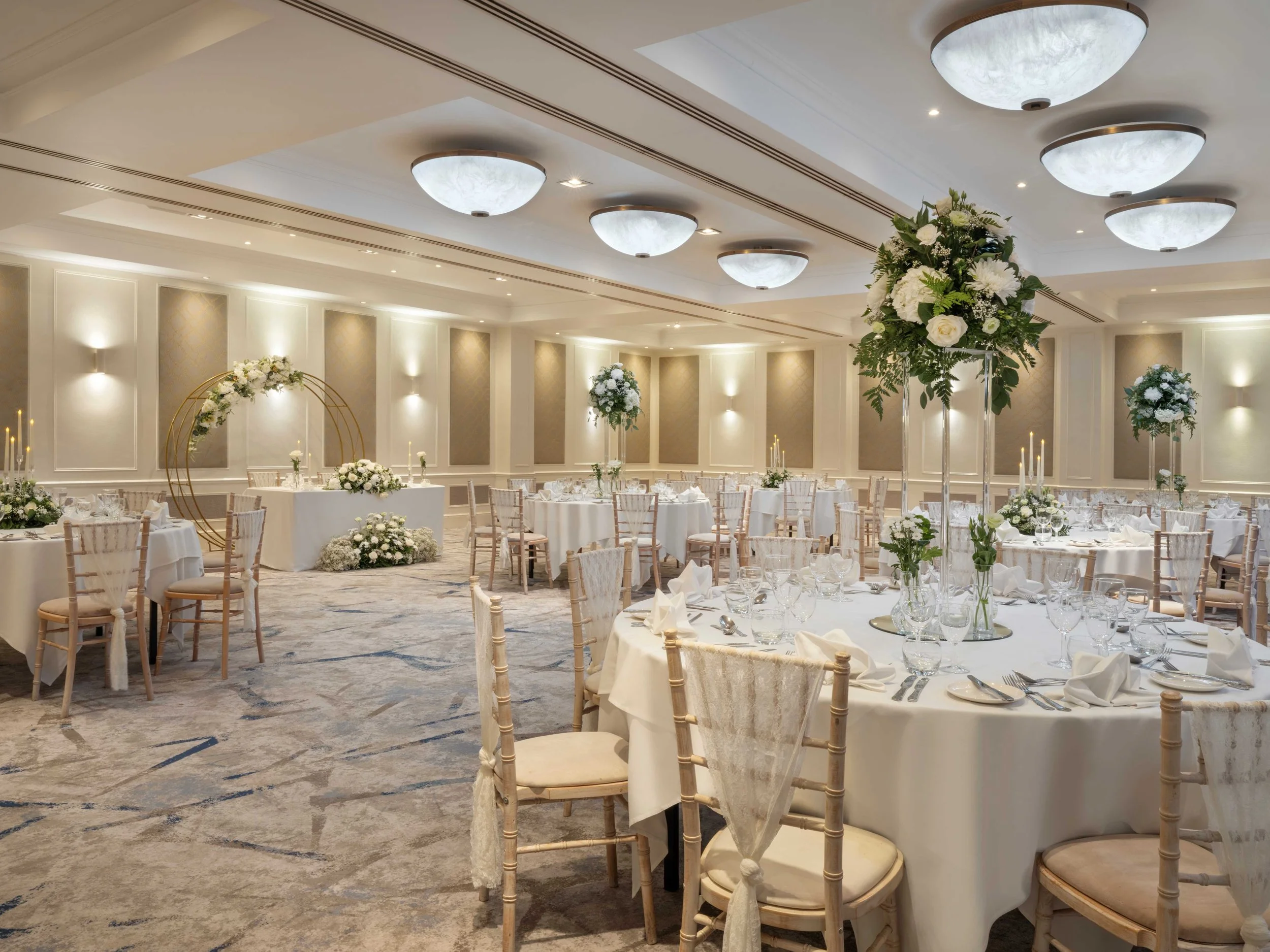 Elegant dining area with a long banquet table set with glassware, candles, and floral arrangements. White chairs surround the table, and a side table on the right holds champagne bottles in an ice bucket and glasses. The room has high ceilings with ornate molding and soft beige walls.