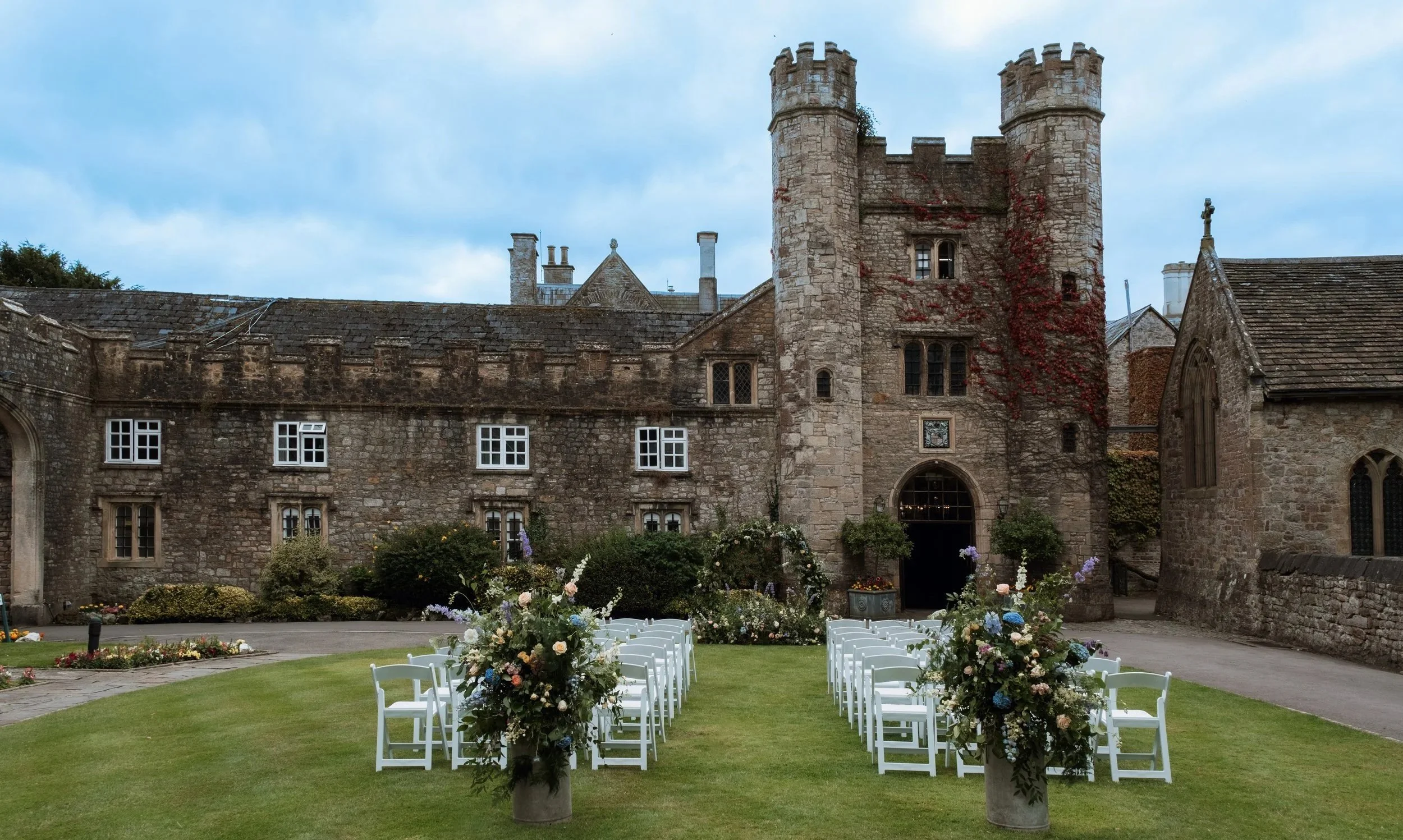 Wedding ceremony setup with white chairs and floral arrangements on the lawn in front of a historic stone castle.