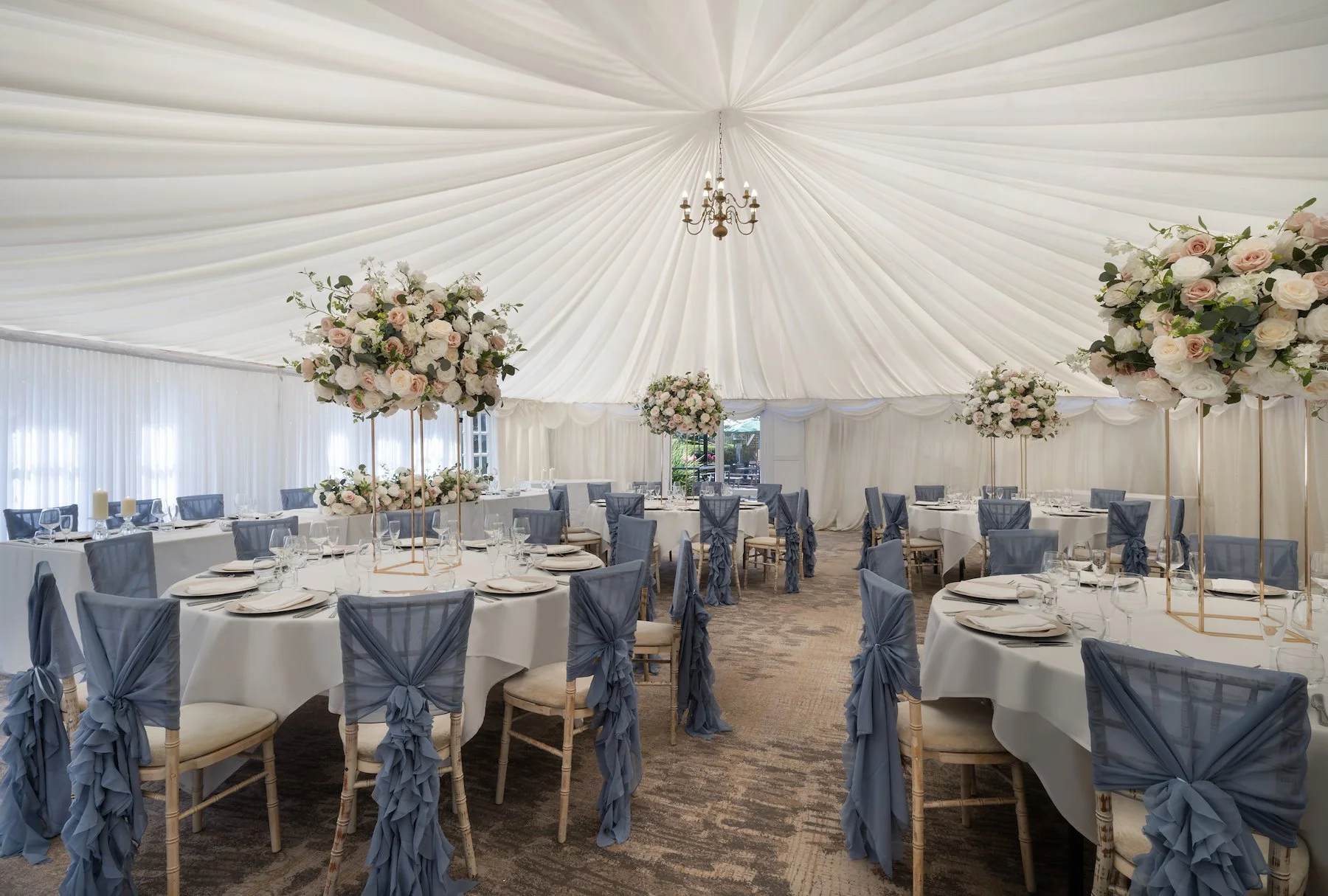 Elegant wedding reception setup inside a tent with round tables, blue cloth chairs, and floral centerpieces on tall gold stands. The tent ceiling is decorated with white fabric and a chandelier.