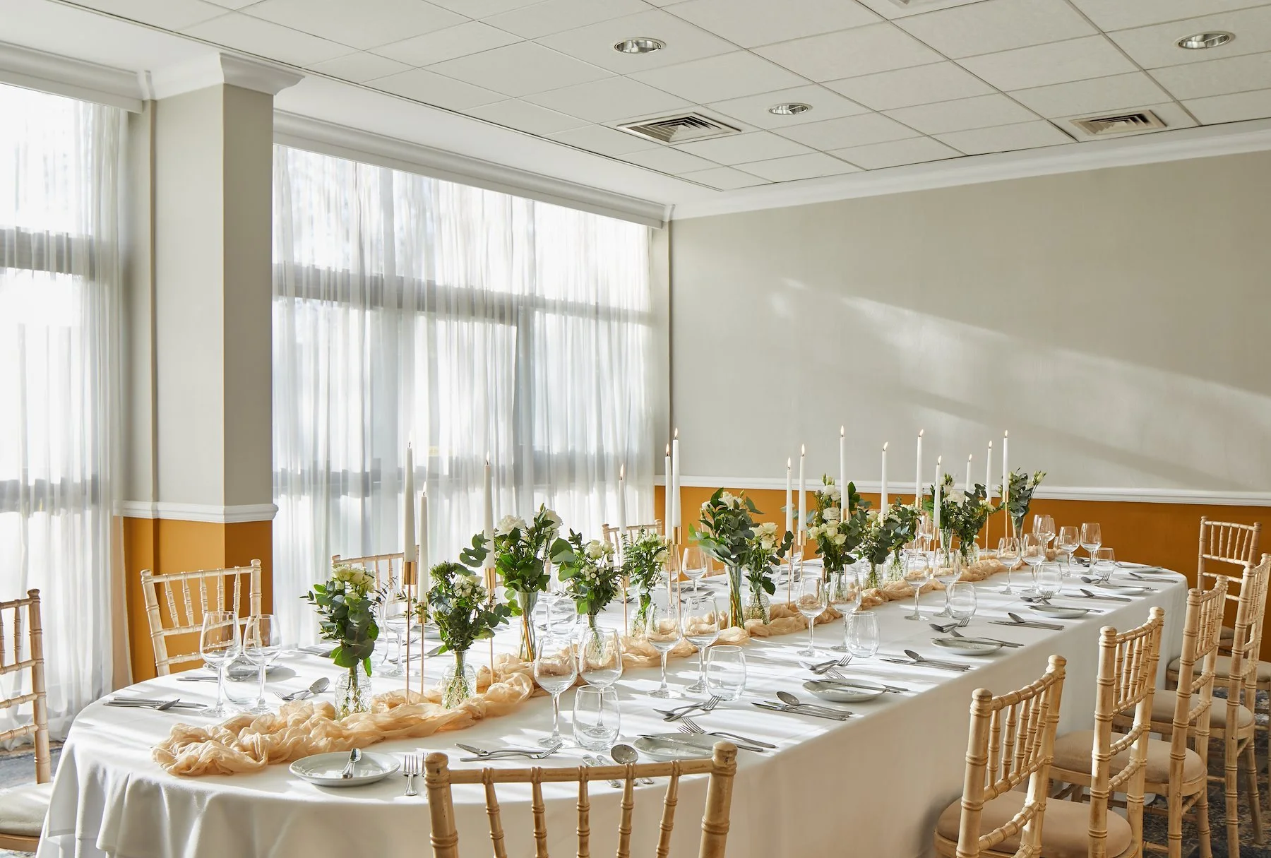 Round banquet table set with glassware, silverware, and floral centerpieces in a well-lit room with large windows and sheer curtains.