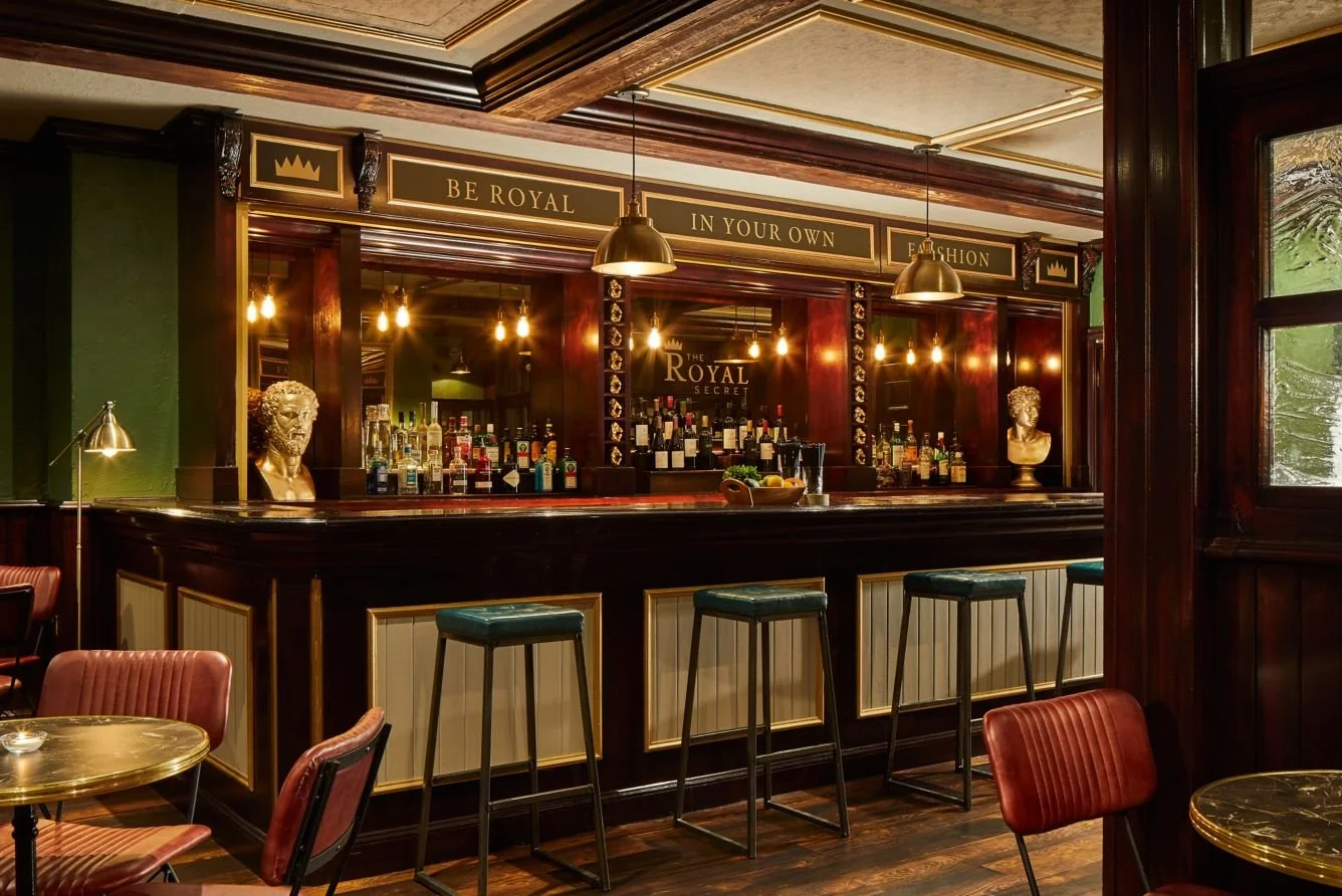 Interior of a vintage-style bar with dark wooden accents, gold decor, bust sculptures, hanging pendant lights, and a polished counter with bar stools.