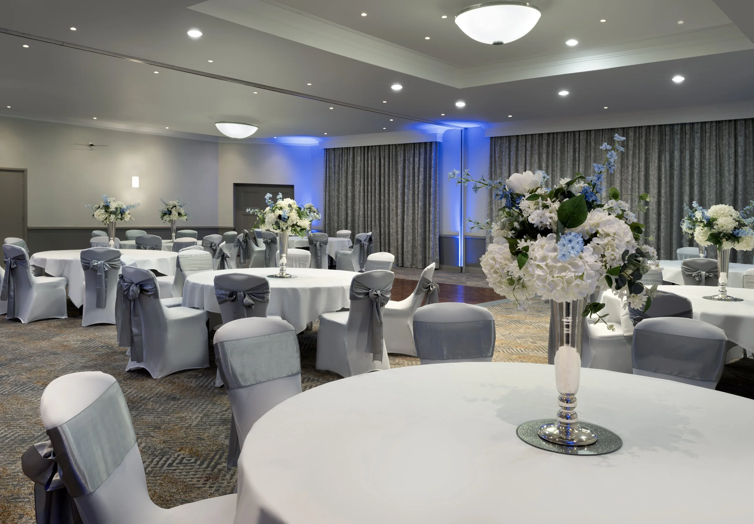 A banquet hall decorated for an event with round tables covered in white tablecloths, each adorned with a tall floral centerpiece featuring white and blue flowers. The chairs are covered with white fabric and tied with gray sashes. The room has a ceiling with multiple recessed lights and large curtains along one wall, illuminated with blue lighting.