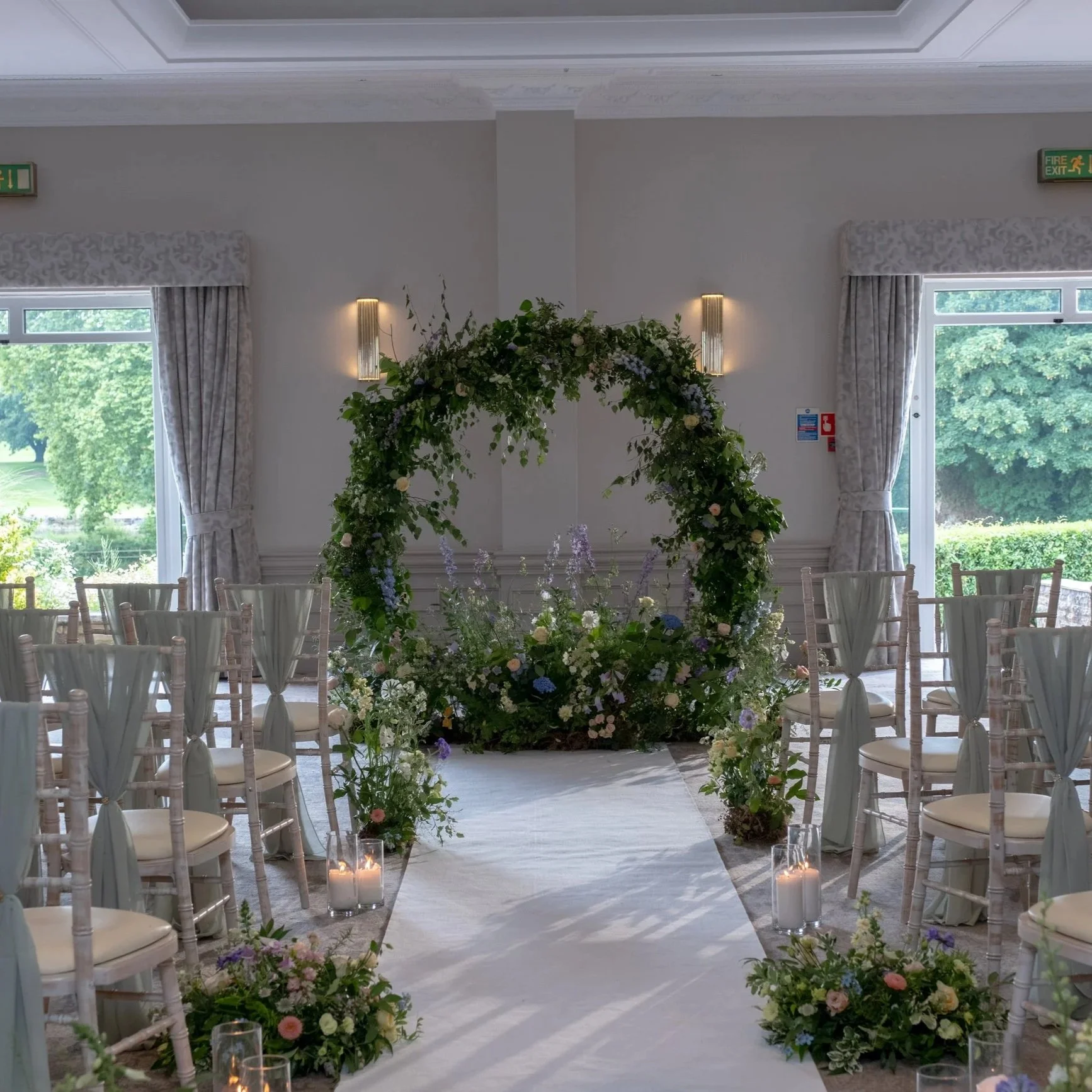 Indoor wedding ceremony setup with floral arch, white chairs with white covers, candles in glass holders, large windows with greenery outside, and soft lighting.