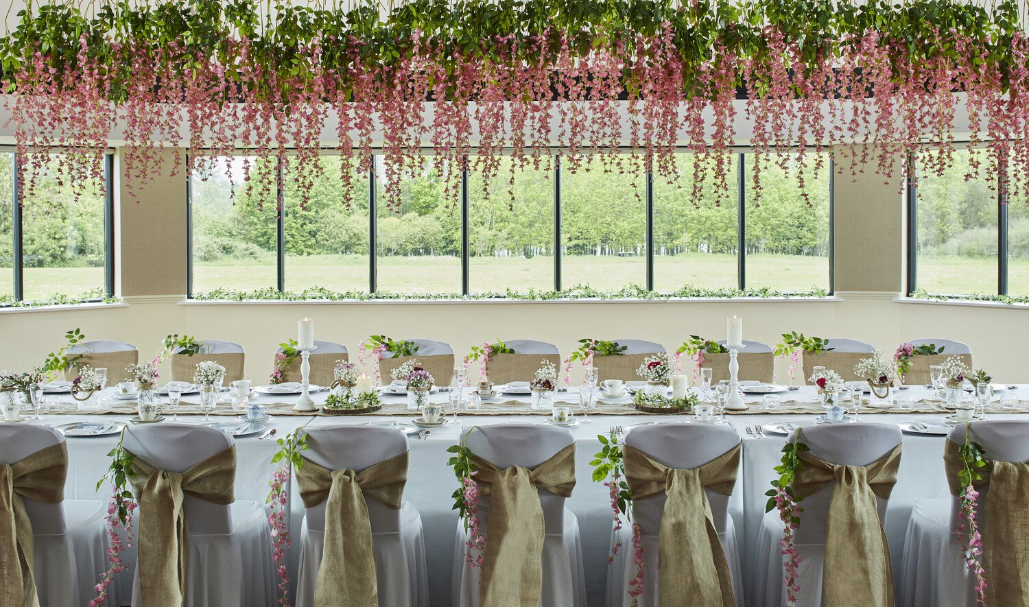 Elegantly decorated banquet hall with a long table set for a celebration, adorned with flowers, candles, and white tableware, featuring beige chairs with burlap bows and a backdrop of large windows revealing a lush green landscape.