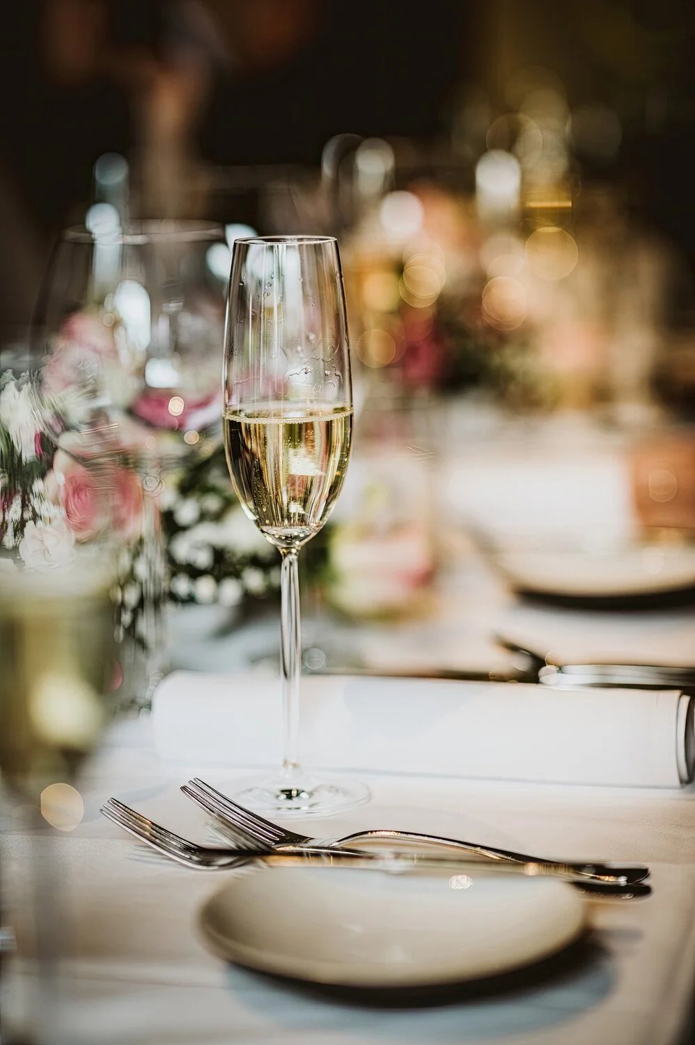 A close-up of a champagne flute filled with sparkling wine on a decorated dining table, with a blurred background of warm lights and floral arrangements.
