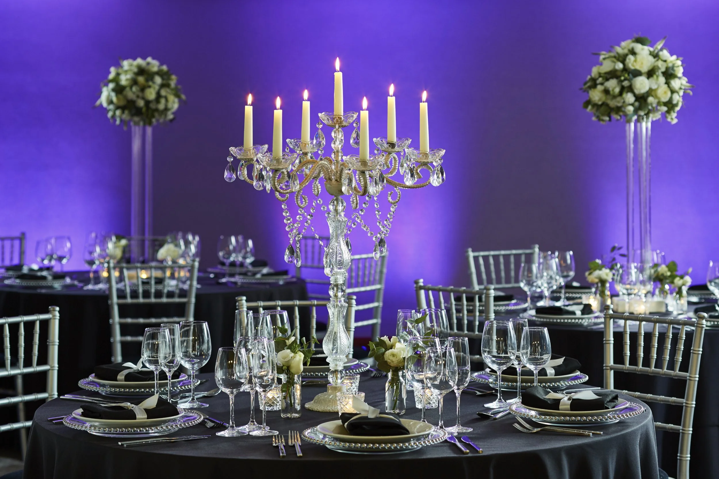 Elegant banquet table with black tablecloths, set with silver chargers, black napkins with white ribbons, and glassware, featuring a tall crystal candelabra with lit candles and hanging crystal ornaments, surrounded by white floral centerpieces, against a purple-lit background.