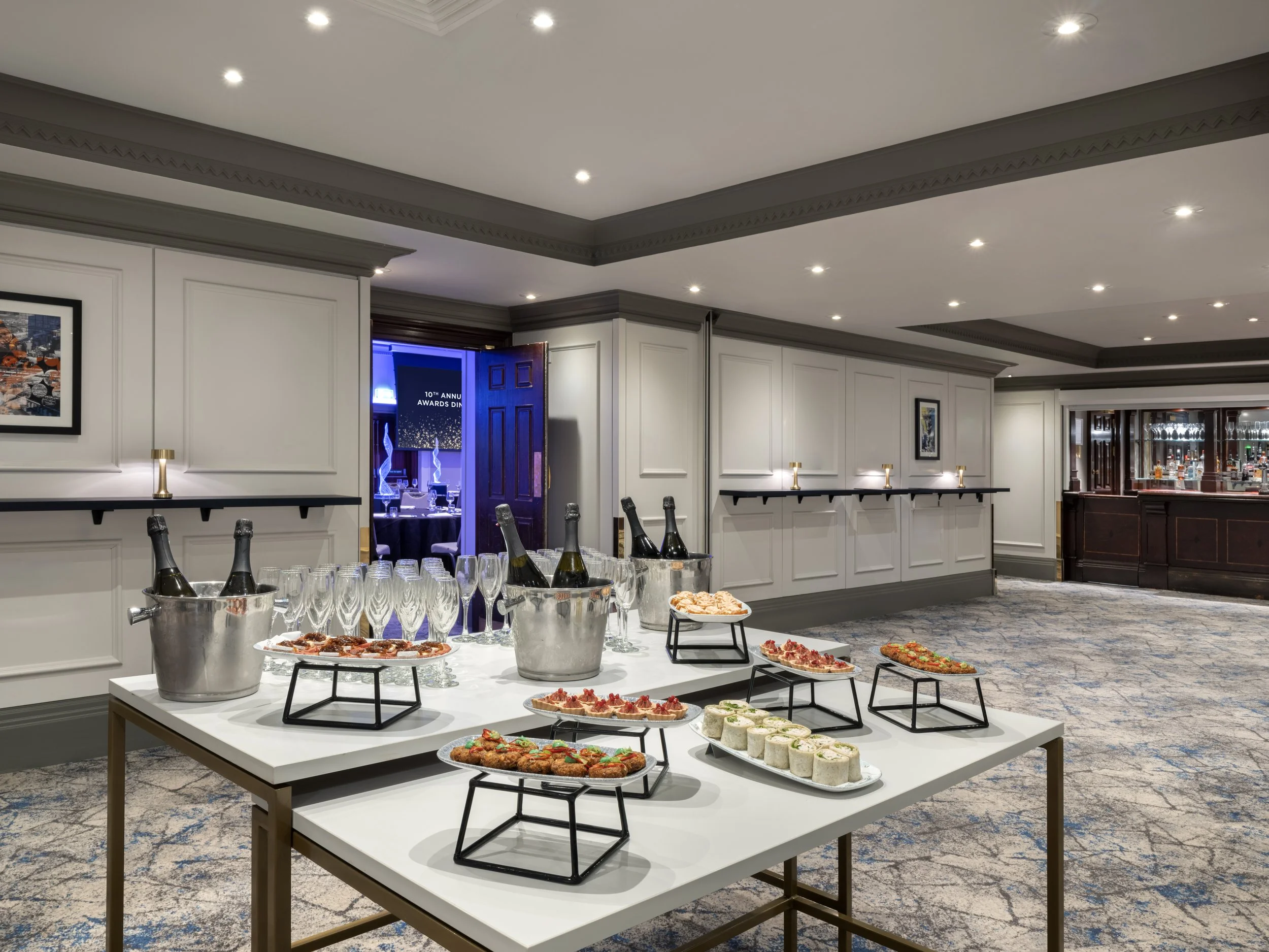 Buffet table with various appetizers and drinks, set in an elegantly decorated room with framed artwork, bar area, and dining space in the background.