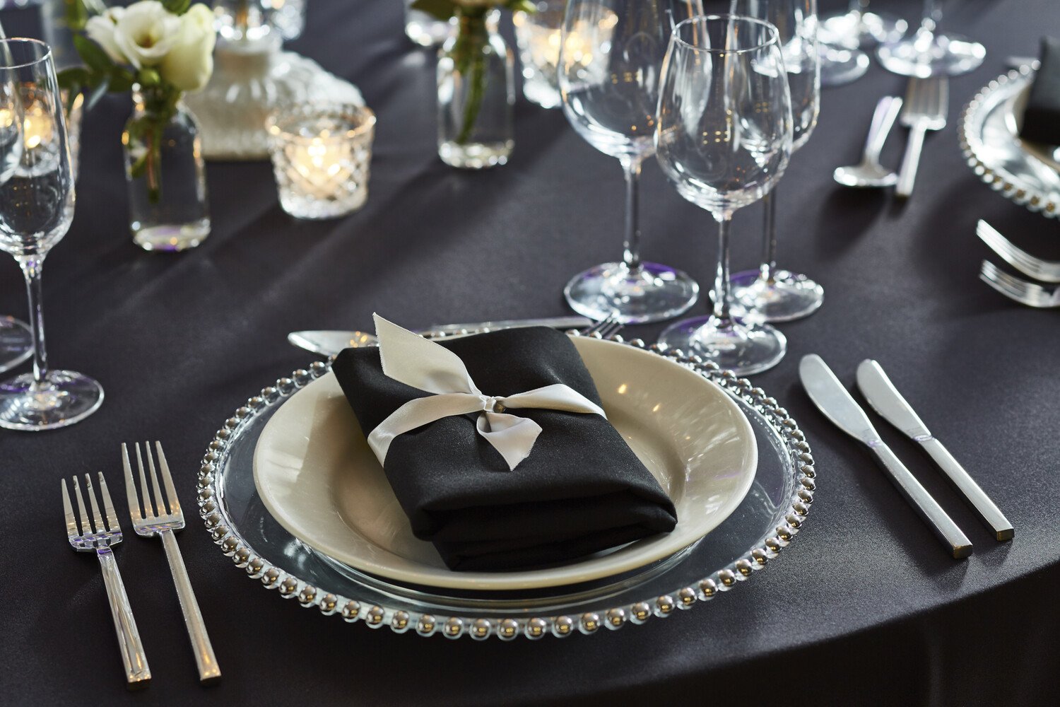 Elegant table setting with black napkin tied with white ribbon on a white plate, surrounded by wine glasses, forks, knives, a candle, and floral arrangement on a black tablecloth.