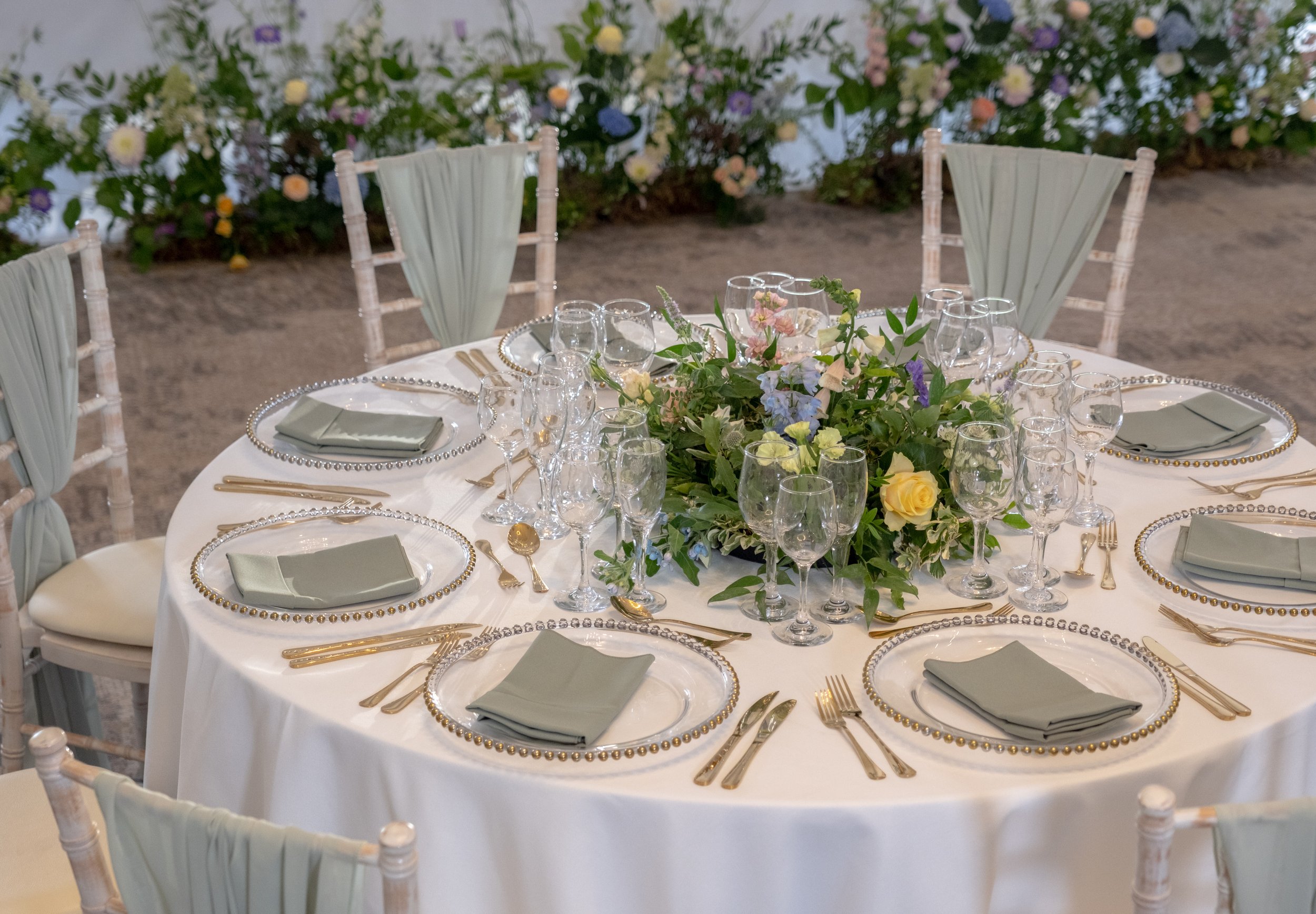 A round banquet table set for a formal event with a white tablecloth, gold flatware, multiple glasses, and light green napkins. The centerpiece features a floral arrangement with pink, yellow, and blue flowers and green foliage. There are wooden chairs with light-colored fabric backrests around the table. In the background, there is a wall decorated with an abundance of flowers and greenery.