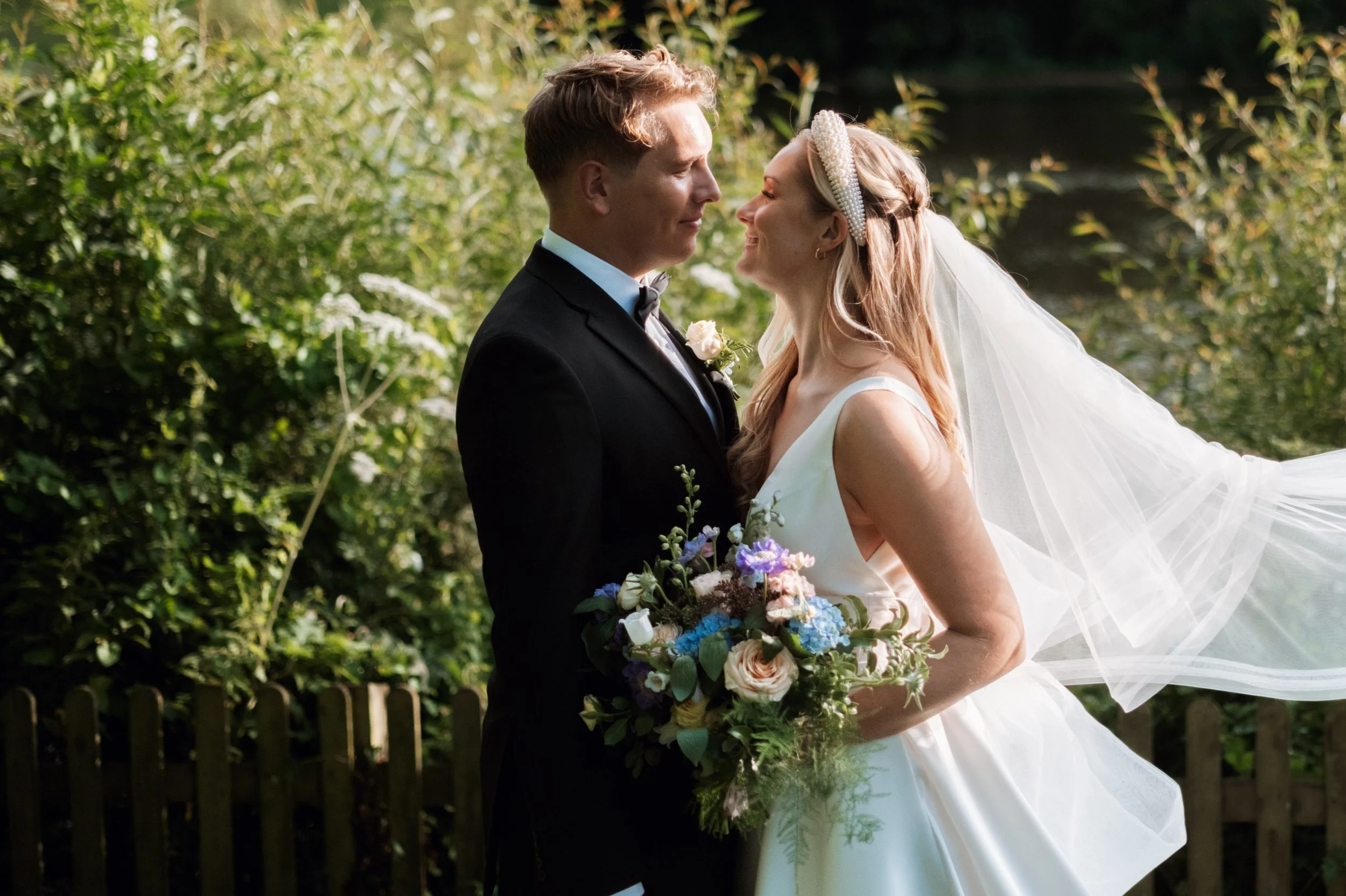 A newlywed couple shares a moment outdoors in the sunlight. The groom in a black tuxedo and the bride in a white wedding gown with a veil and pearl headband stand close, smiling at each other. The bride holds a large bouquet of flowers, and lush greenery surrounds them.