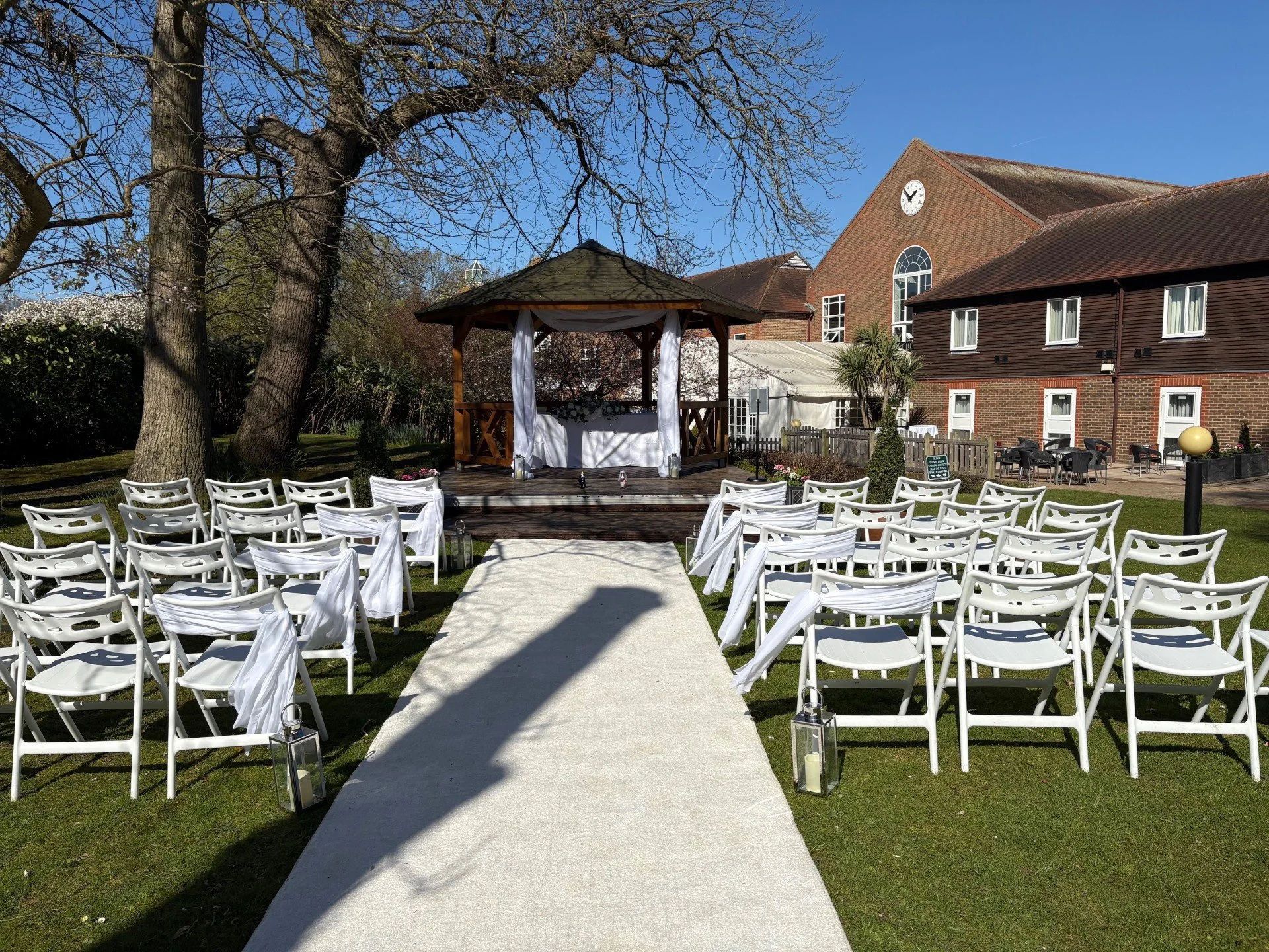 Outdoor wedding ceremony setup with white chairs on both sides of a white aisle runner, a gazebo decorated with white fabric, trees, and a brick building in the background under a clear blue sky.