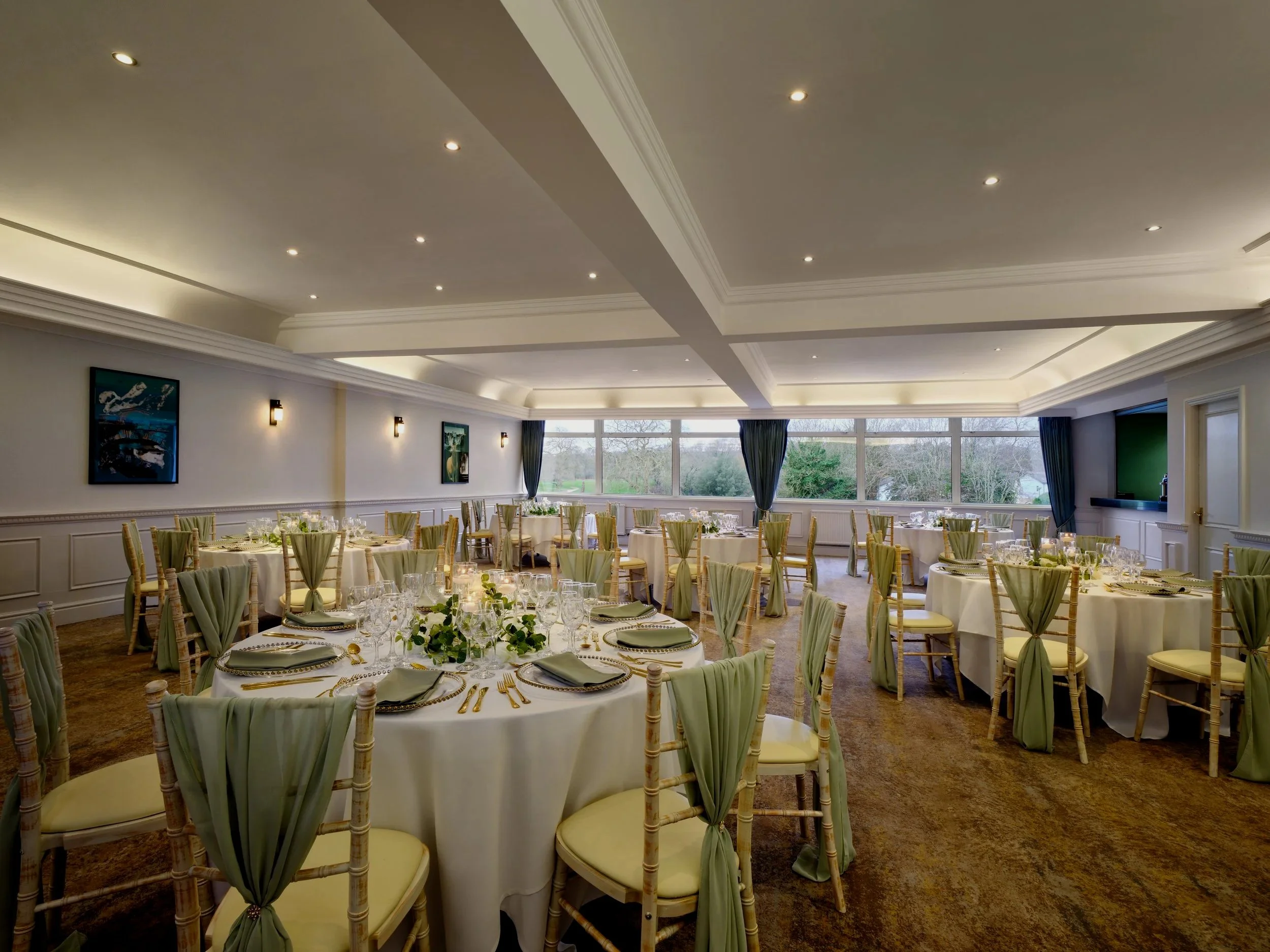 An elegantly decorated banquet hall with round tables covered in white tablecloths, set with glassware, silverware, and napkins, surrounded by yellow chairs with green sashes. Large windows with blue curtains reveal a scenic outdoor view. The ceiling has decorative molding and is illuminated by recessed lights.