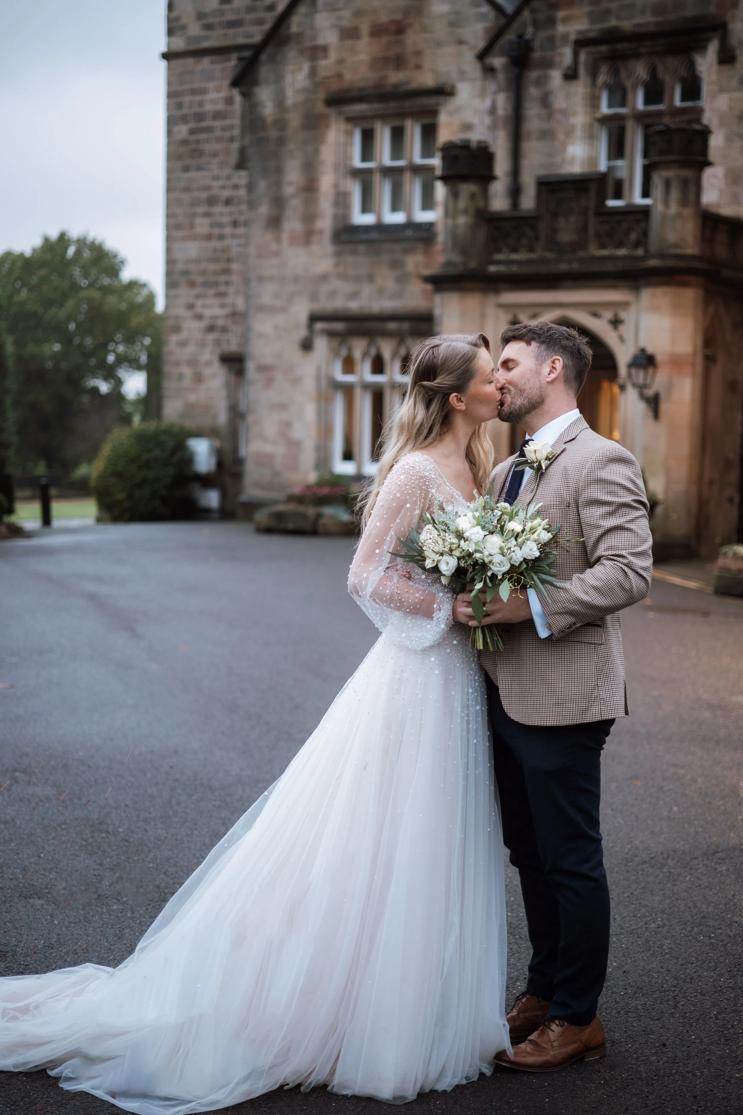 A bride and groom kissing outside a historic stone building, with the bride holding a bouquet of white roses and greenery, and the bride wearing a white wedding gown with a long train.