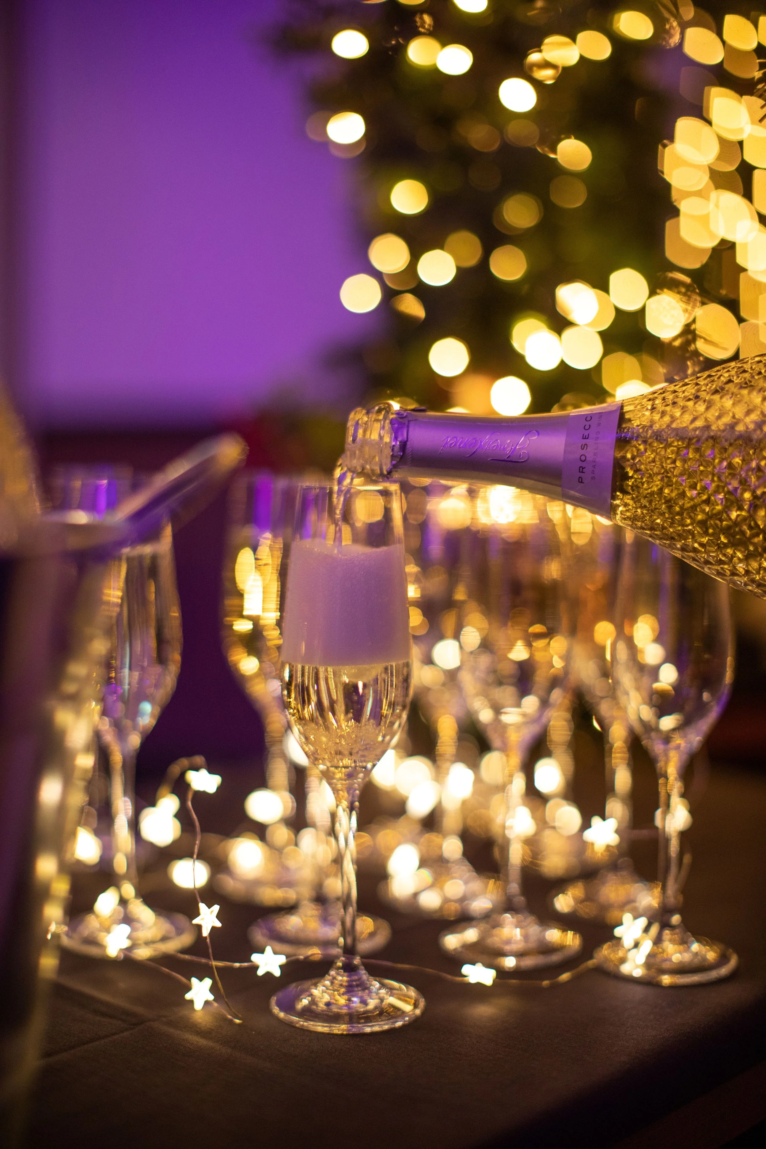 A row of champagne glasses with a champagne bottle labeled 'Prosecco' pouring into one glass, set against a background of blurred yellow and purple lights, resembling a festive celebration.
