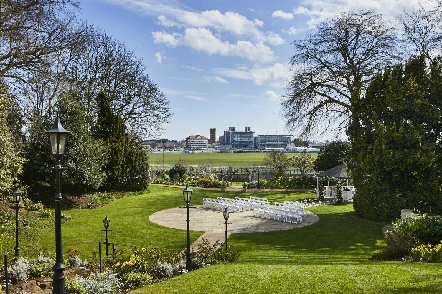 A garden with green grass, flowerbeds, and black lampposts, overlooking a racecourse with a large grandstand in the distance under a partly cloudy sky.