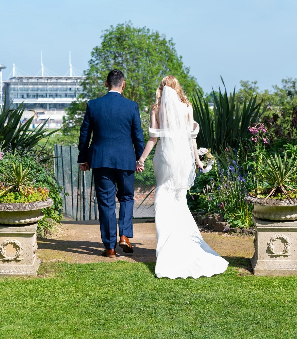 A bride and groom walking hand in hand outdoors on a sunny day, surrounded by greenery and flowers, with a large building in the background.