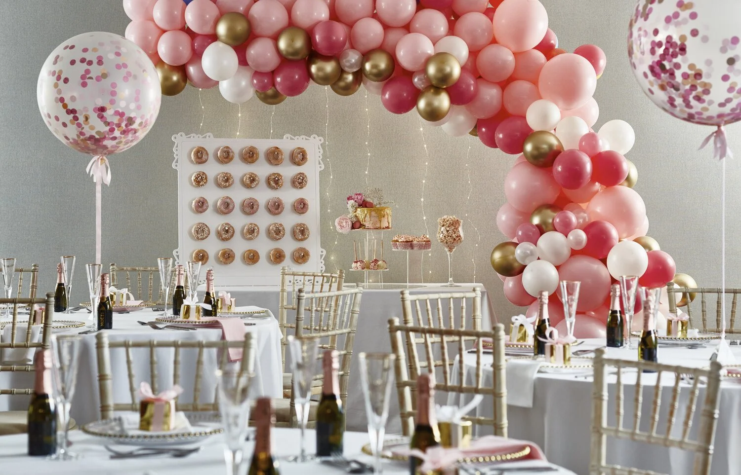 A decorated event space with pink, white, and gold balloons forming an arch, two large confetti-filled balloons flanking the scene, and tables set with champagne bottles, glasses, and small gift boxes. There is a dessert display with donuts, cakes, and pink confections.