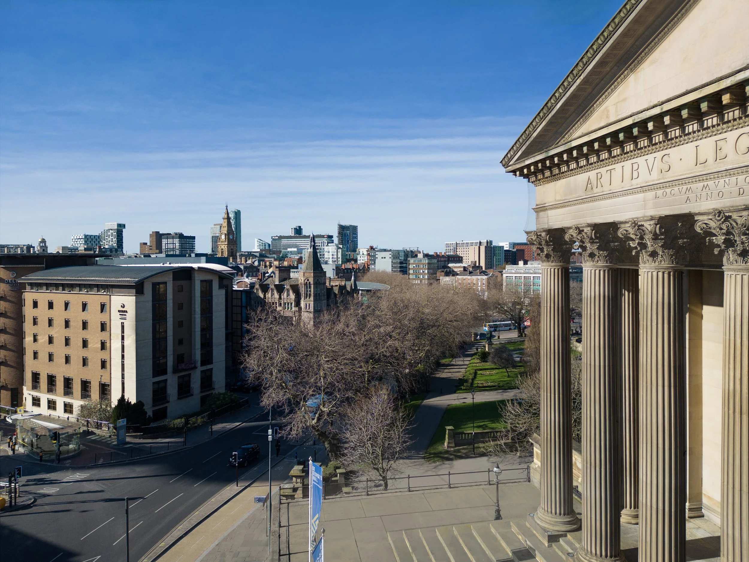 View of a city skyline with modern and historic buildings, seen from the steps of a neoclassical building with large columns and Latin inscriptions, under a clear blue sky.