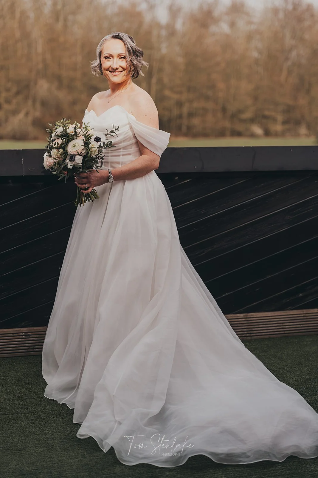 A woman in a white wedding dress holding a bouquet standing outdoors on a balcony with trees in the background.