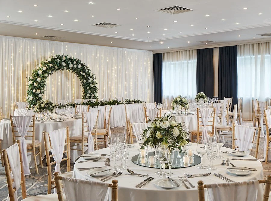 Elegant wedding reception setup with round tables, white tablecloths, floral centerpieces, and a floral arch in the background.