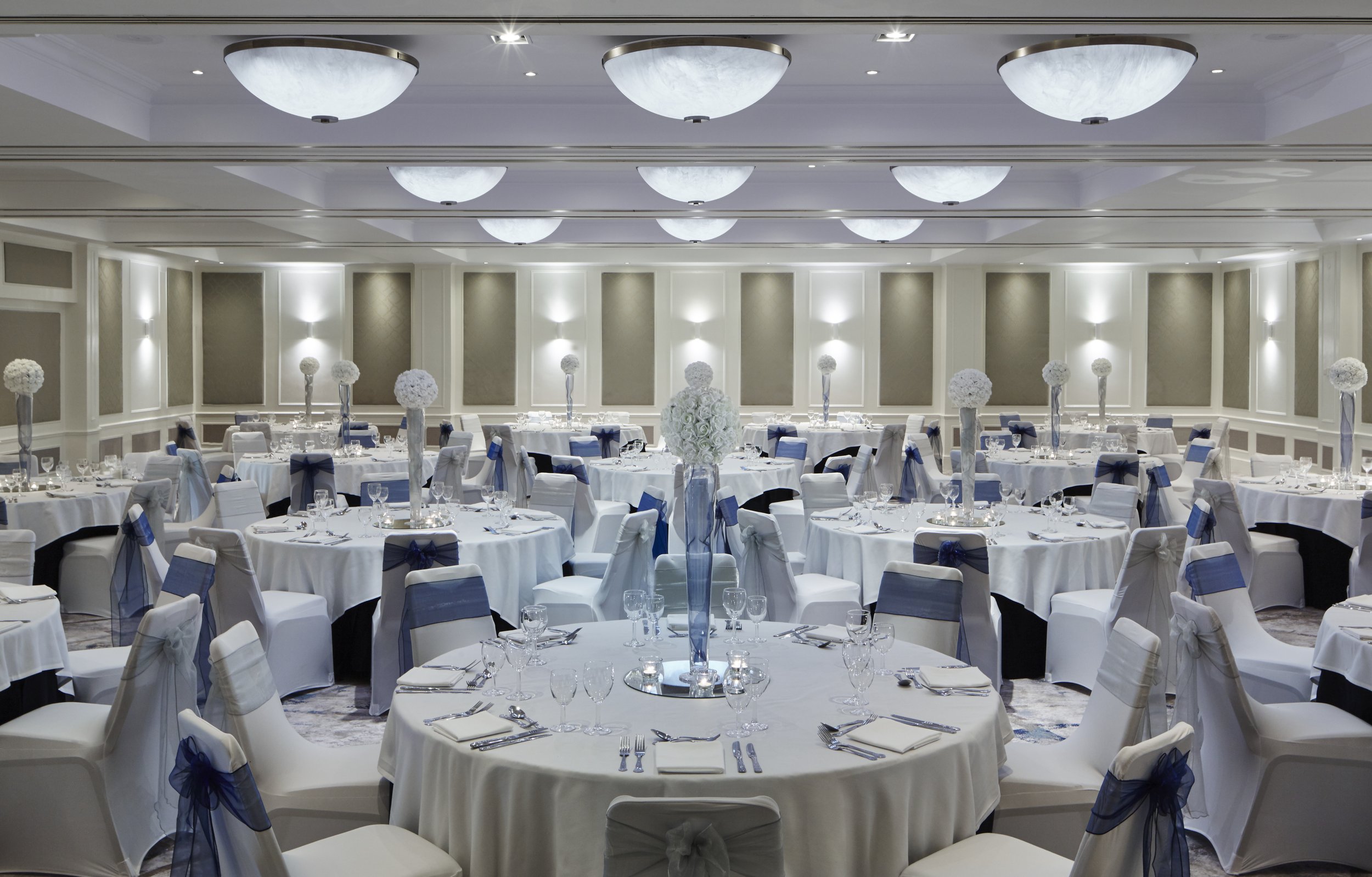 Elegant banquet hall decorated for a formal event with round tables covered in white tablecloths, adorned with tall floral centerpieces, and surrounded by white chairs with navy blue ribbons. The room has soft, white lighting and paneled walls.