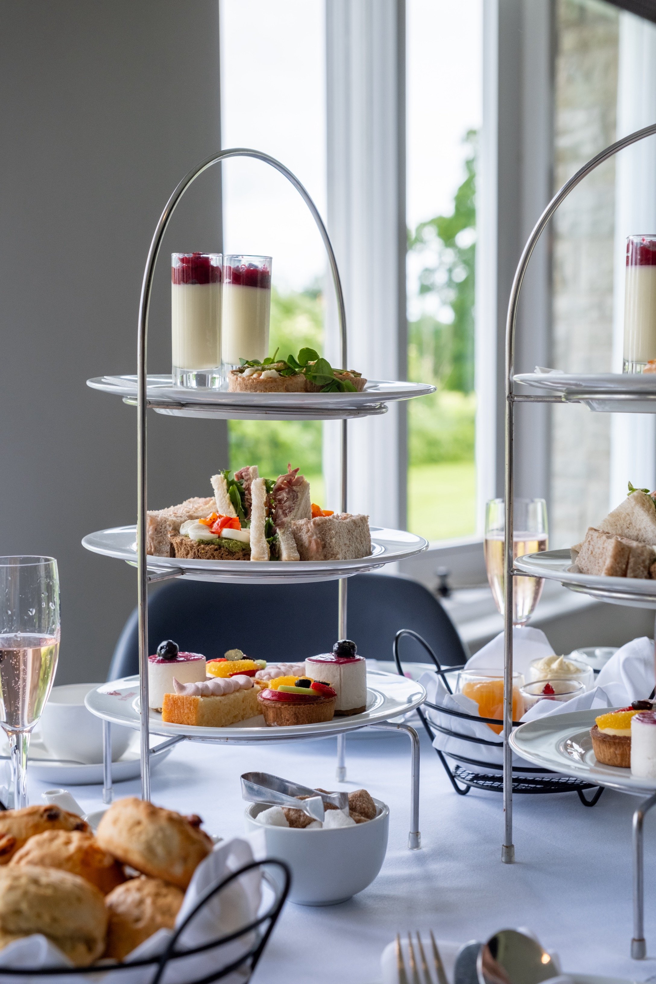 A tiered serving tray with assorted desserts, including panna cotta with berry topping, finger sandwiches, and small cakes, set on a white tablecloth near a window with a view of greenery outside.