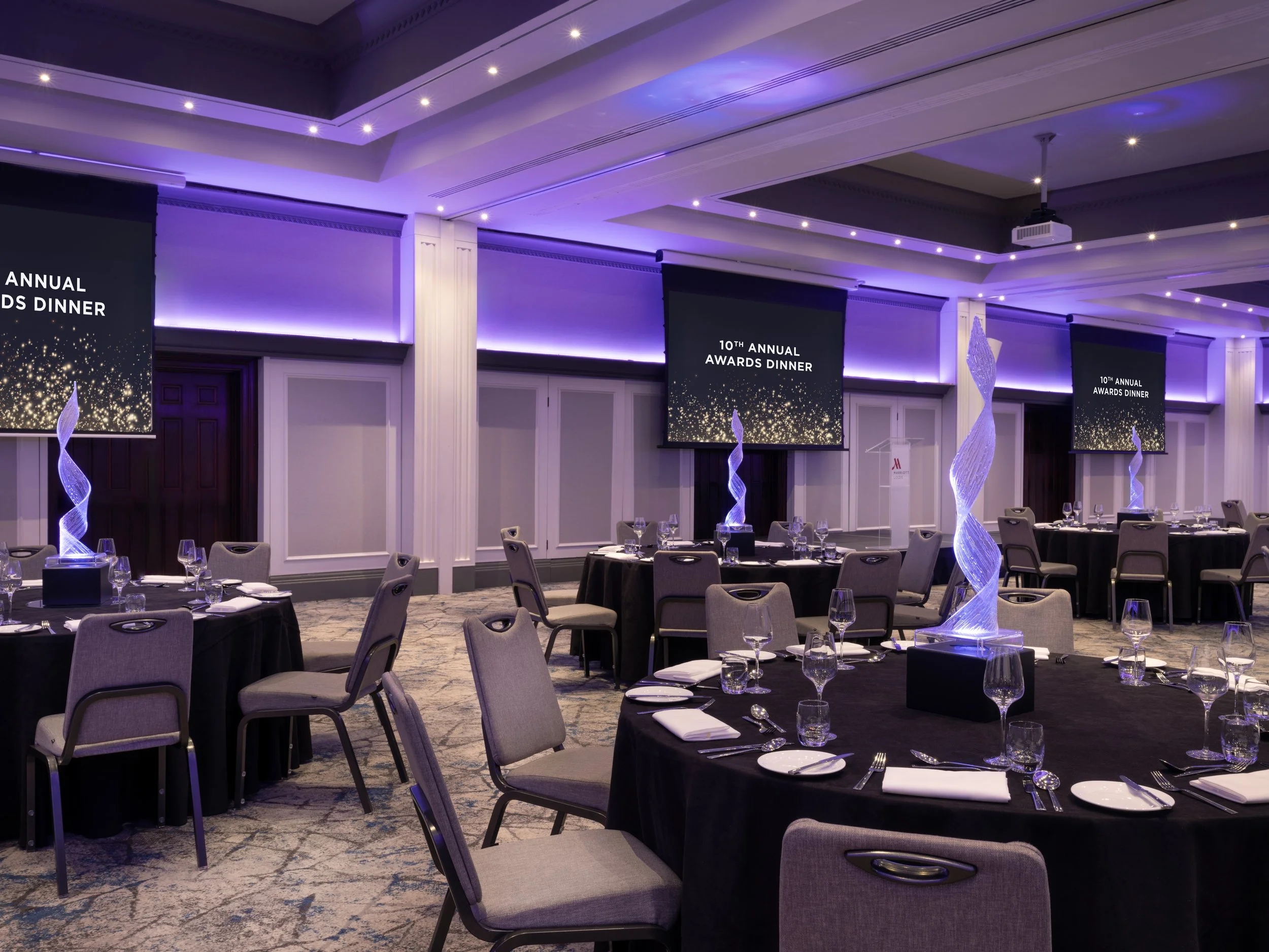 Elegant banquet hall set up for an awards dinner with round tables, black tablecloths, and place settings. Large screens display '10th Annual Awards Dinner' with a sparkling gold and black design. Purple ambient lighting and decorative twisting light sculptures enhance the ambiance.